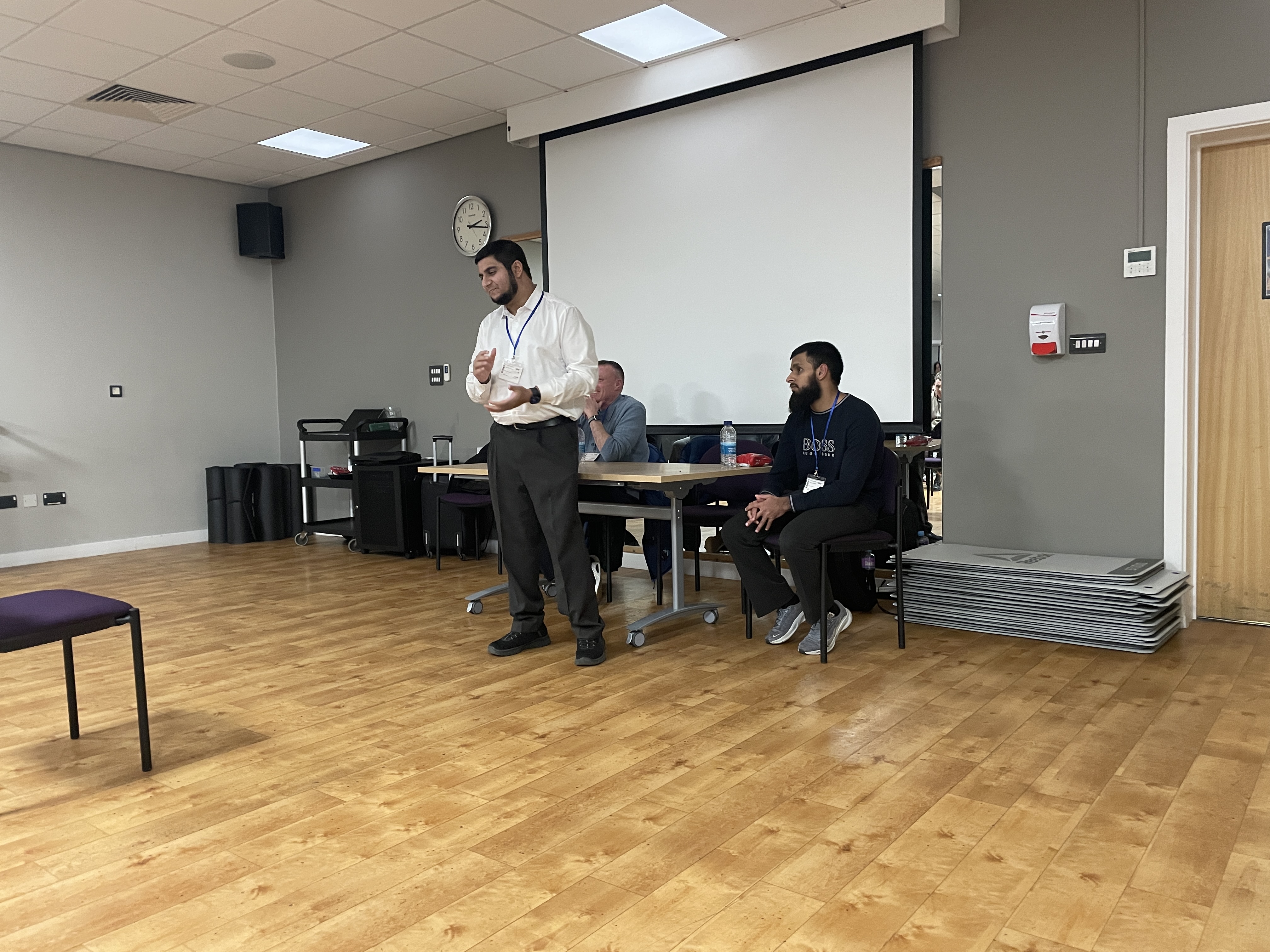 Mo stands at the front of the room facing his audience. Barry and Eesa are sat at a table behind him.