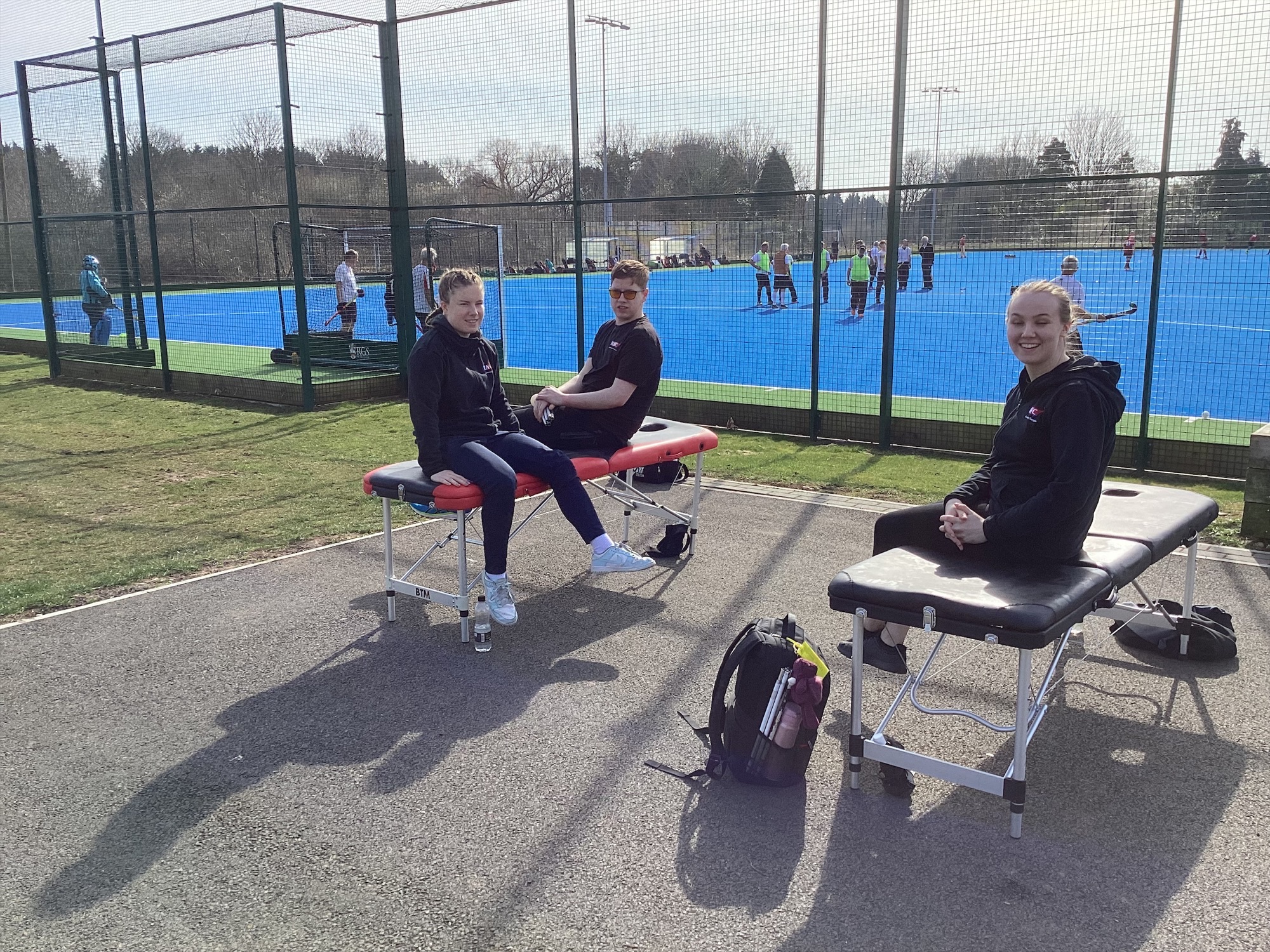 three students sitting on portable massage couches. A game of hockey is going on the courts behind them.