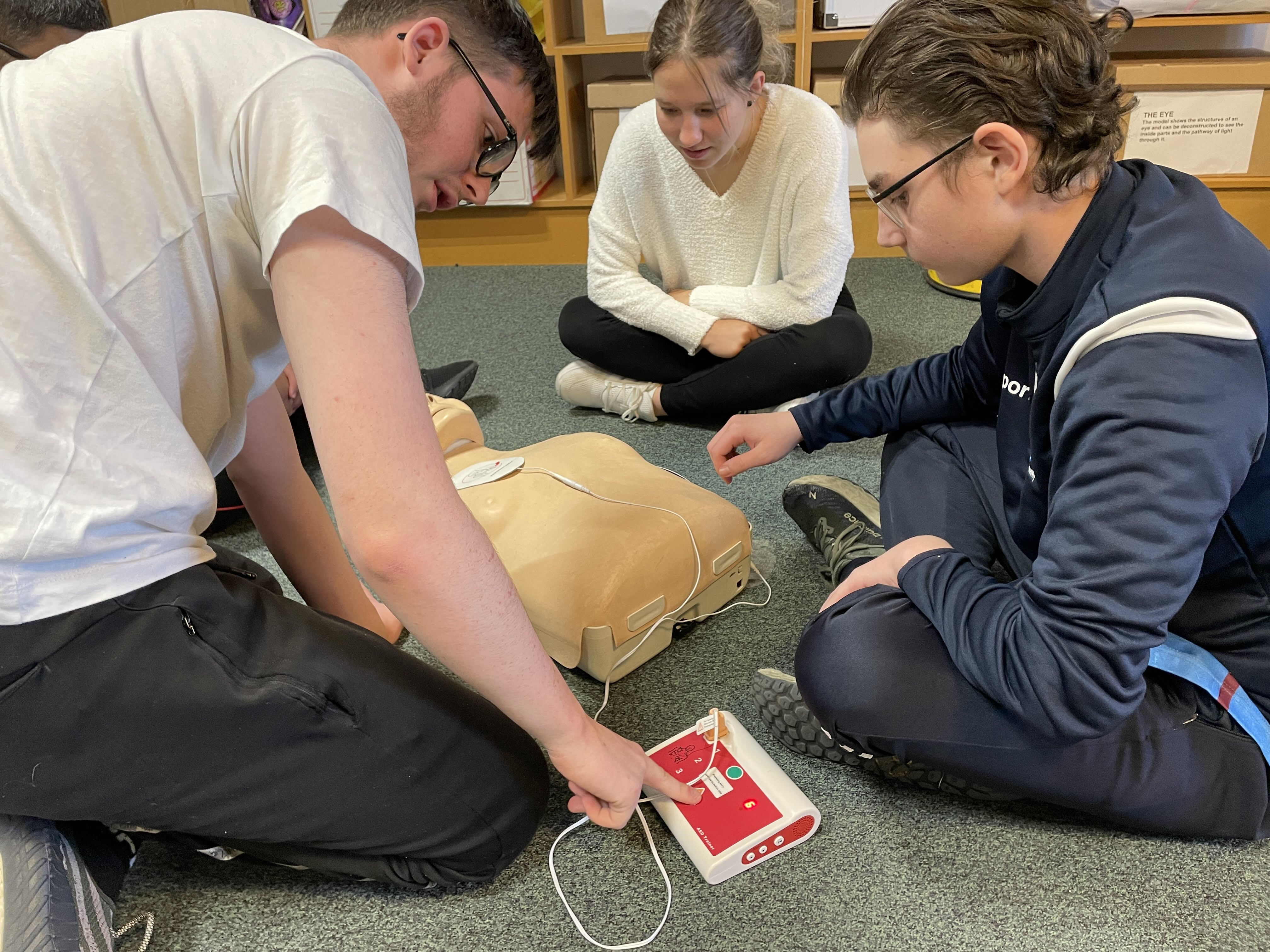 3 people gathered around a resusci Annie doll, the student on the left is about to press a button on the defibrillator