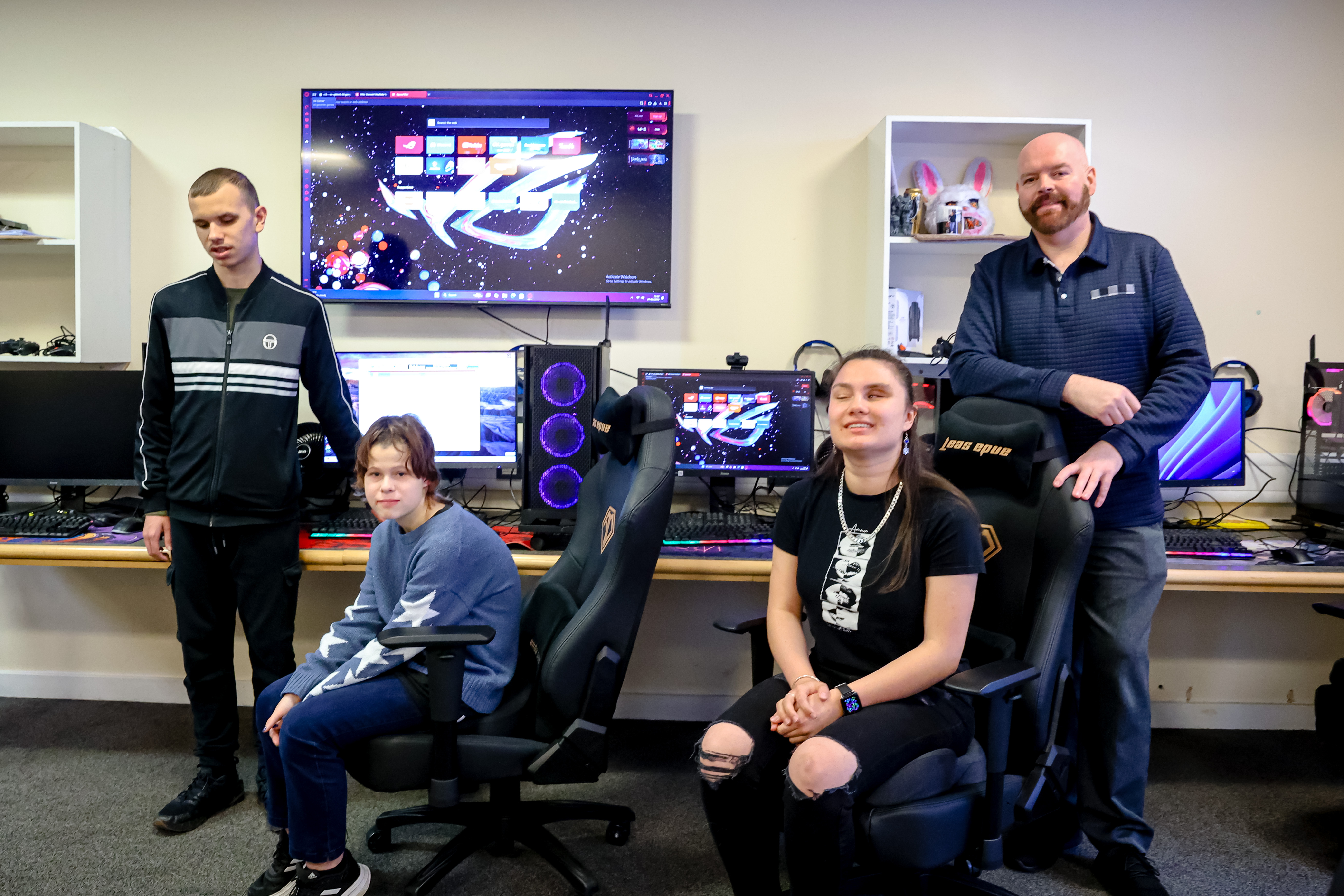the 3 students and their teacher stand, or sit in gaming chairs in front of gaming computers with bright colours on the screens