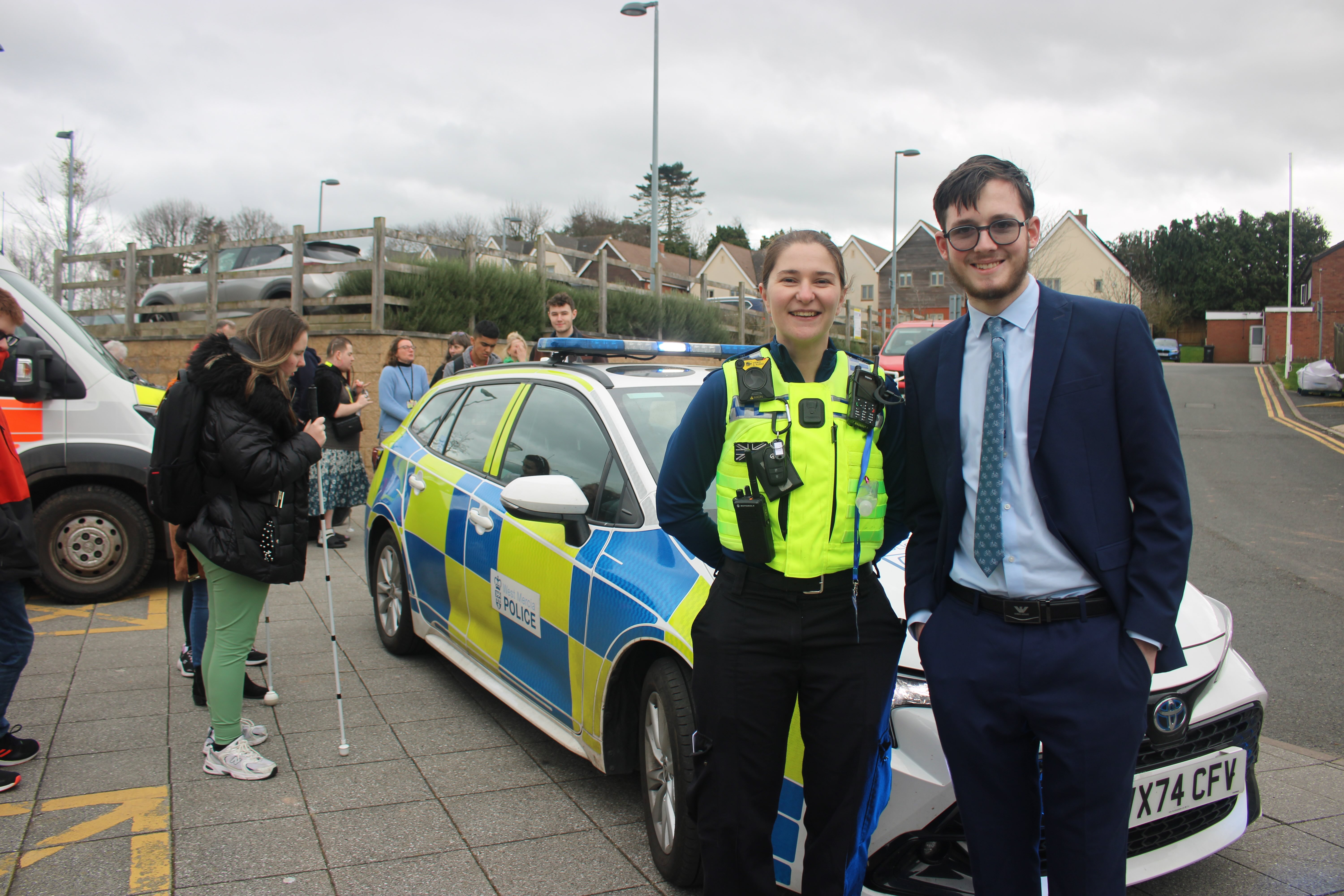 Elliot stands with the Police Woman in front of the police vehicle - students are milling around in the background