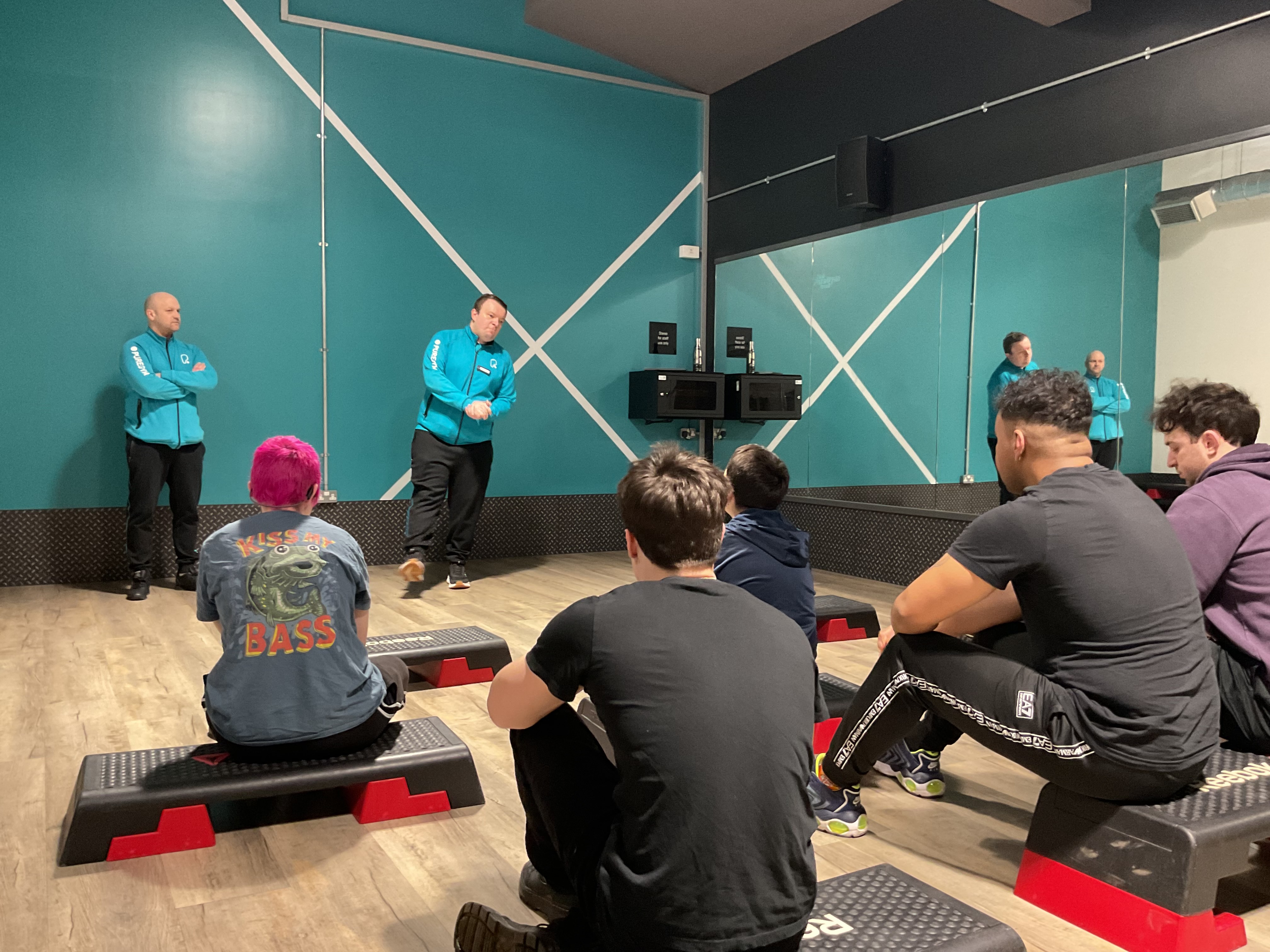 Jack stands in front of the group who are sitting on steps in a fitness studio in front of him