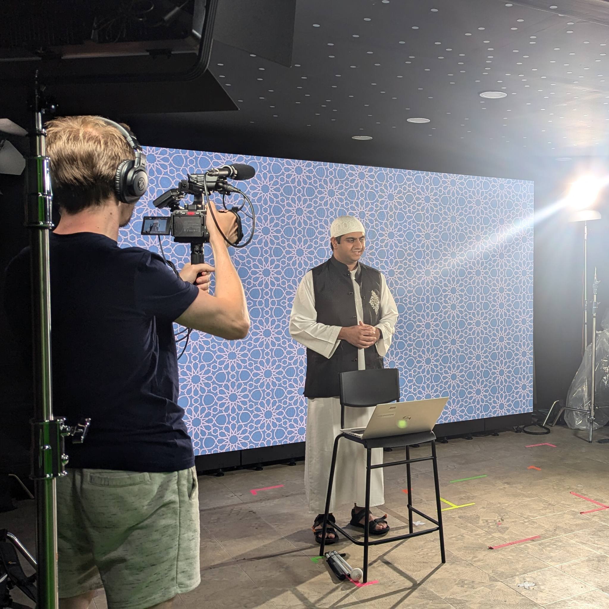 Mo stands in front of a large screen with Islamic art on it. There is a cameraman with camera filming Mo