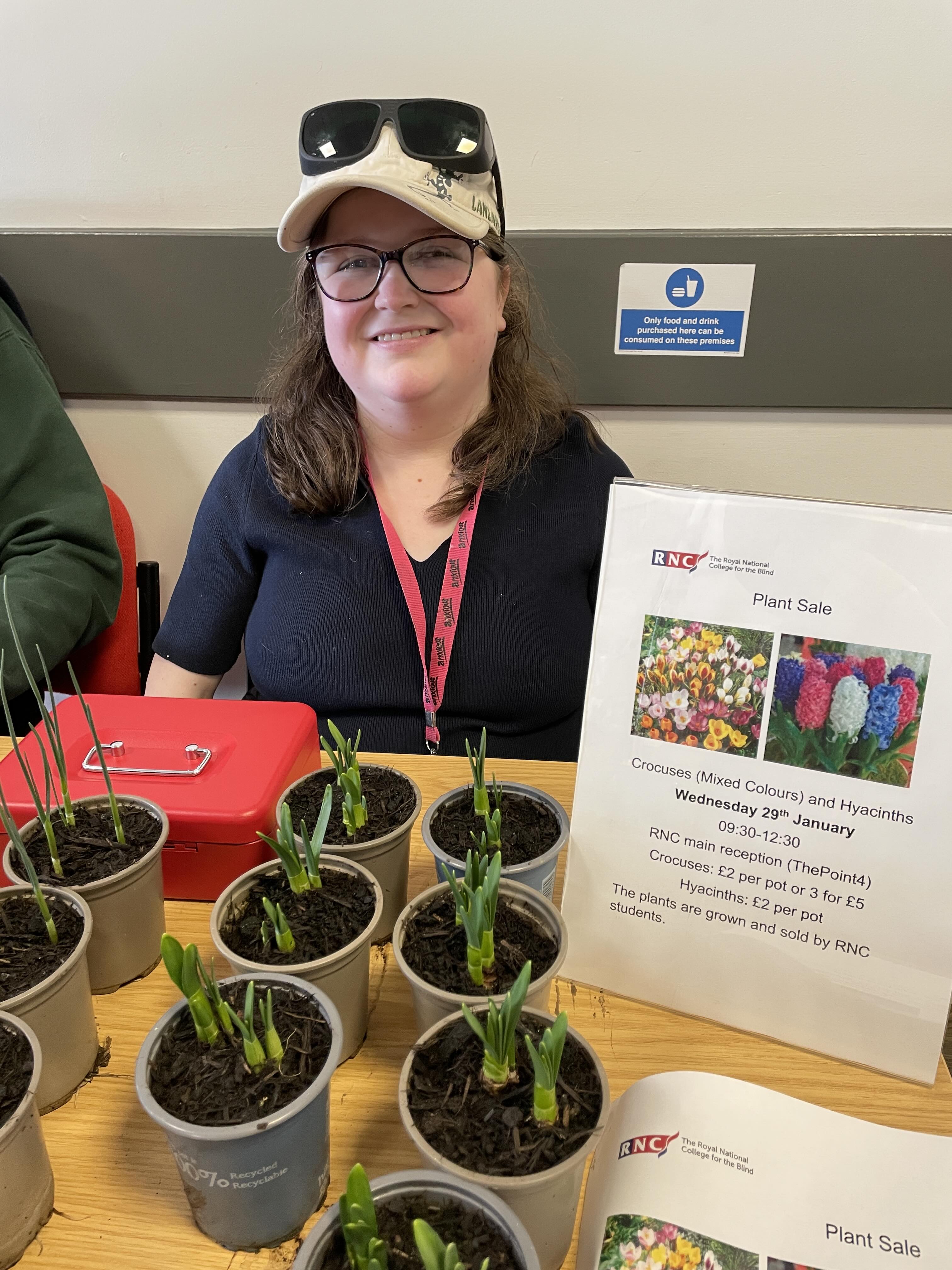 Katie sits behind a table with potted plants in front of her and a cash box and a poster advertising the plant sale