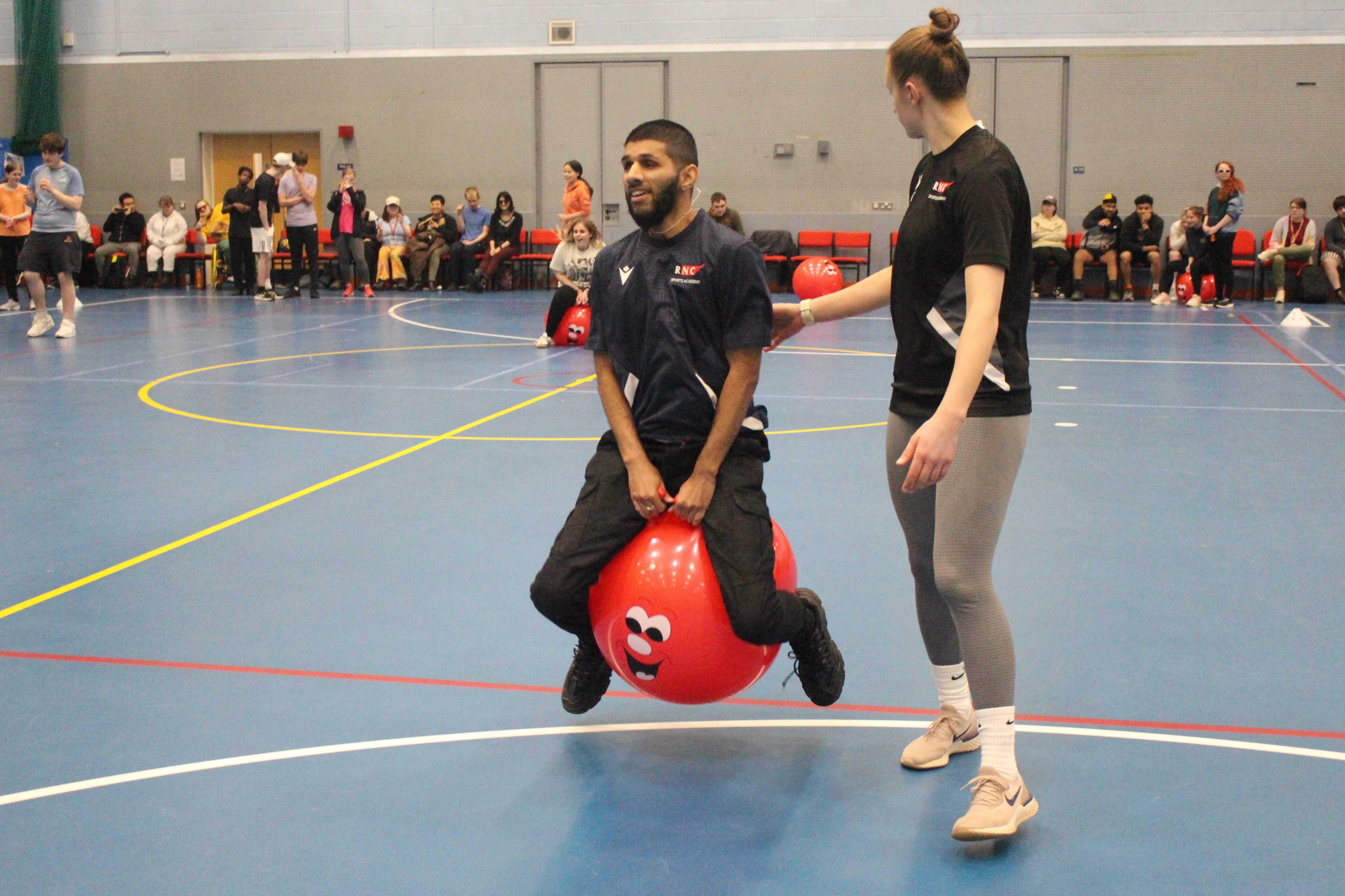 make student racing on a red space hopper - he's bouncing in the air - a female member of staff is next to him for safety