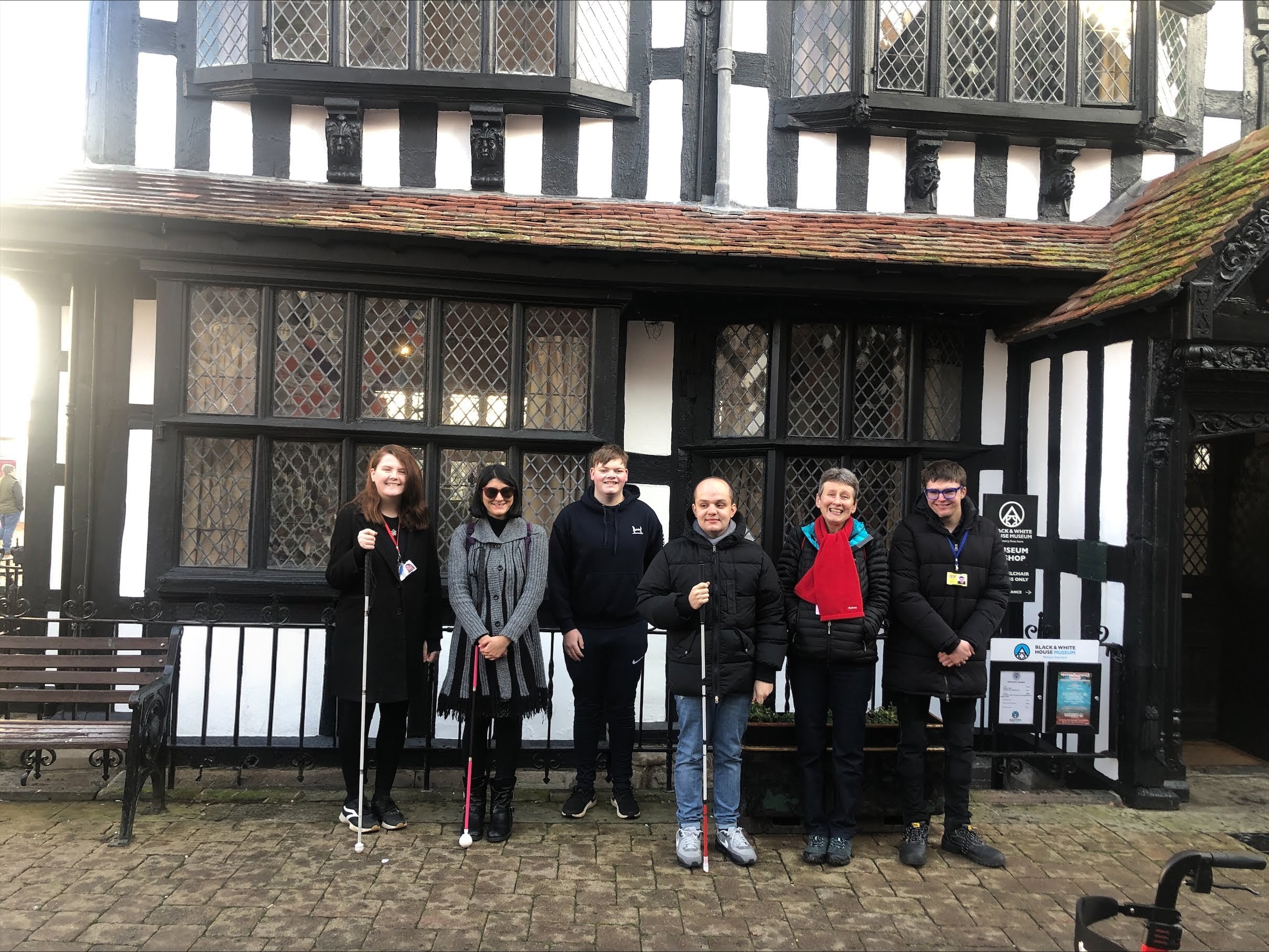 A group of 6 students, volunteers and staff stand in a line in front of the Old House. The entrance is to the right and their are leaded windows visible behind the group.