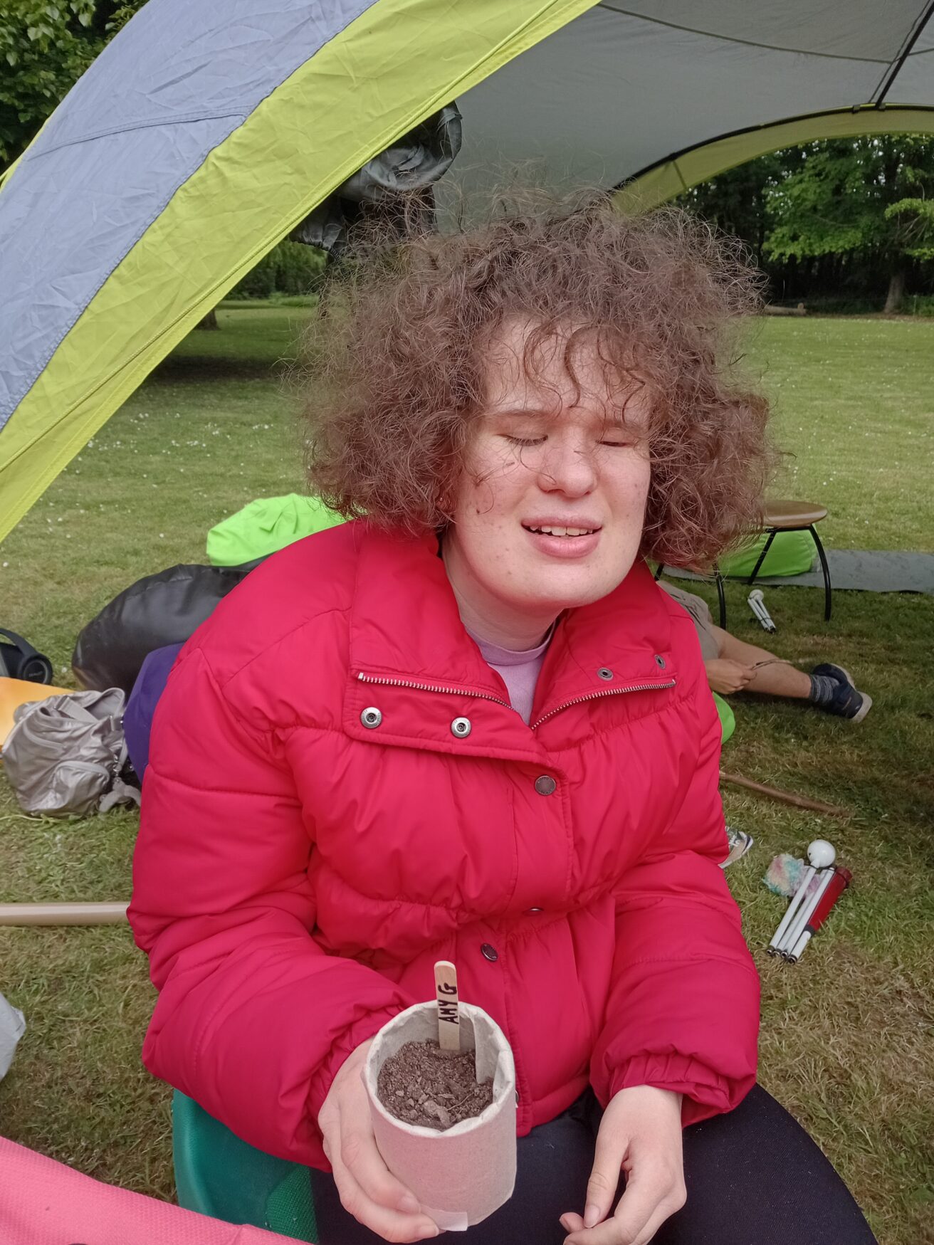 Amy sitting on the orchard, holding up a pot which she has planted a sunflower in