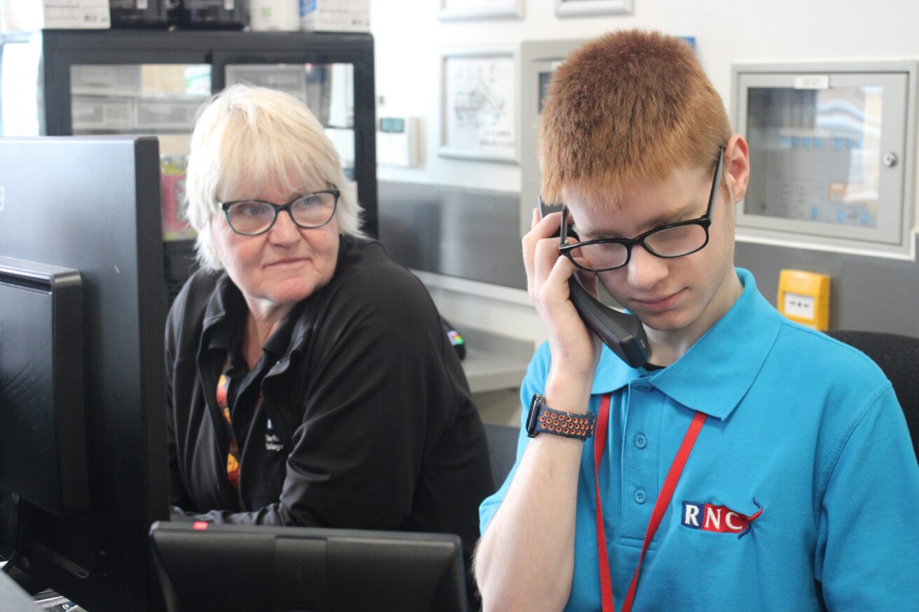 Oli sitting behind reception holding a telephone receiver to his ear. Julie is watching on in a supportive way