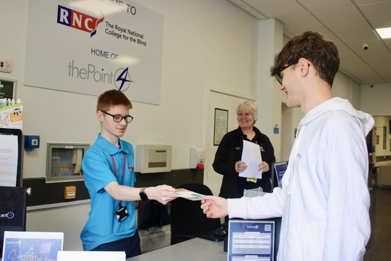 Oli is stood up behind the reception desk handing a leaflet to a visitor - Julie is looking on smiling