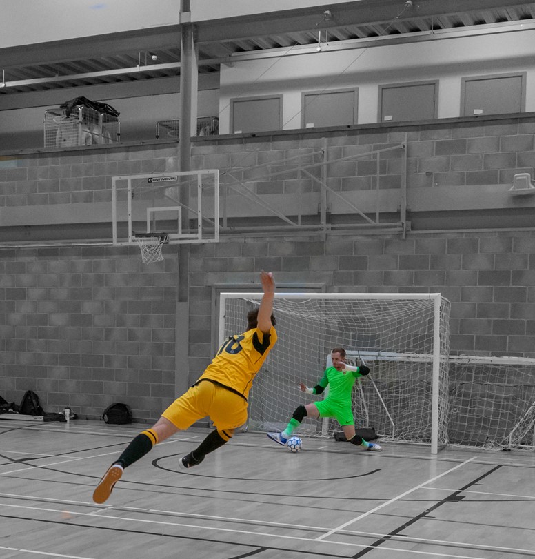 Photo of a futsal match, with a player running toward to the goal with the ball, and the goalie preparing to defend.