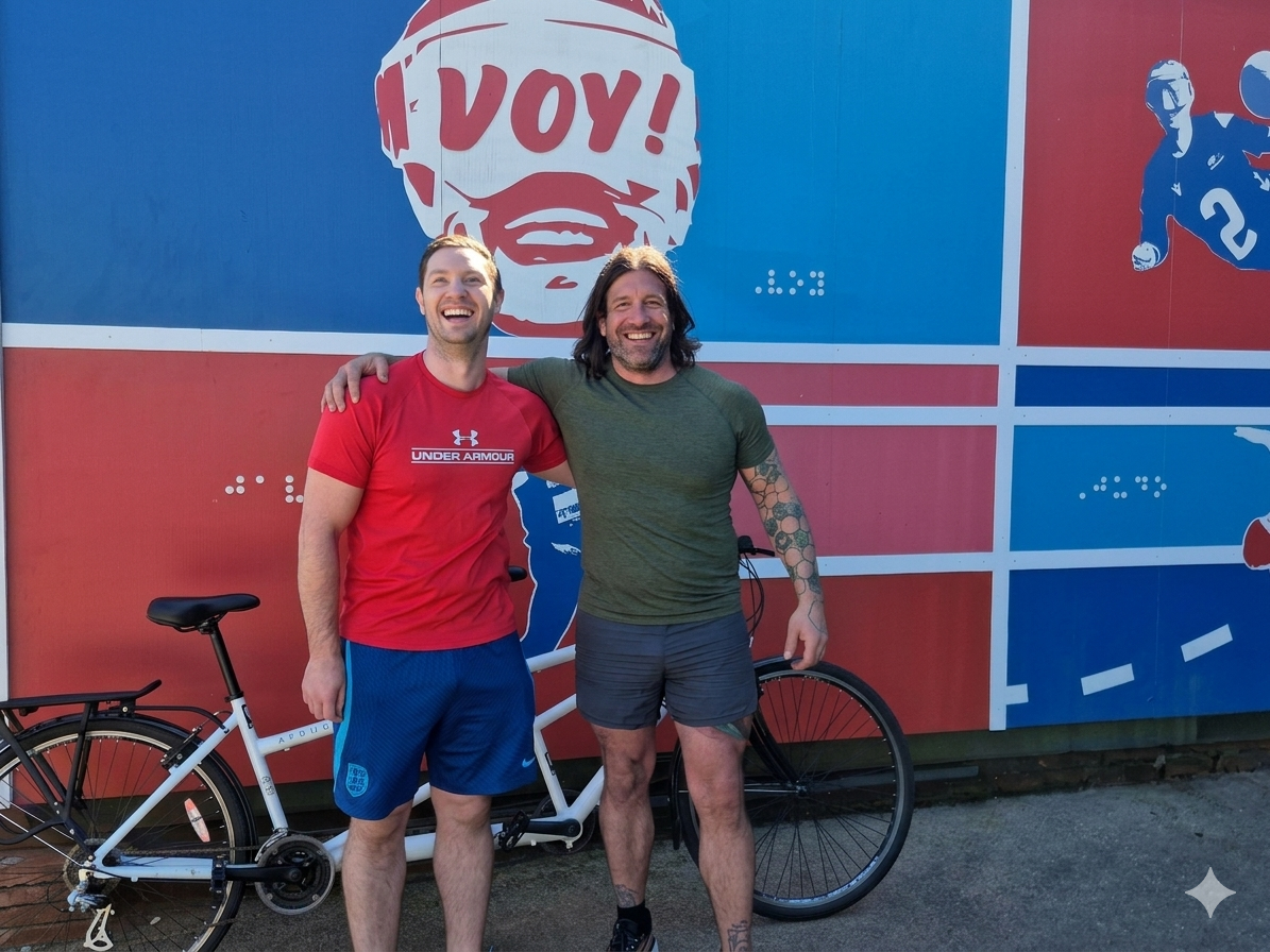 Owen a Gareth gyda'u beic tandem yn sefyll o flaen celf graffiti myfyrwyr sy'n cynnwys Owen yn gwisgo mwgwd pêl-droed i bobl ddall uwch eu pennau