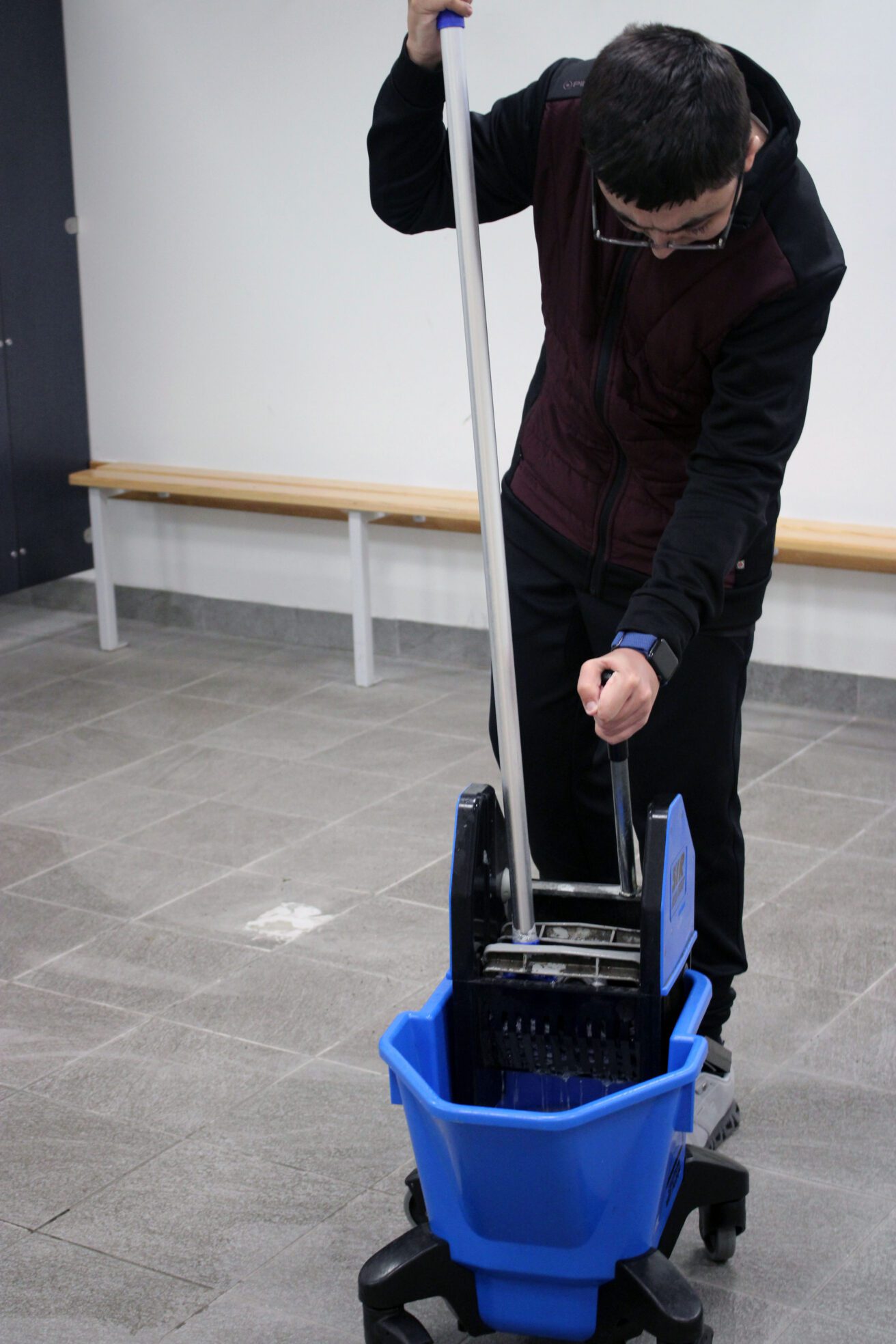 Emilio holds the mop in one hand while pulling the handle on the blue mop bucket to squeeze out the water.
