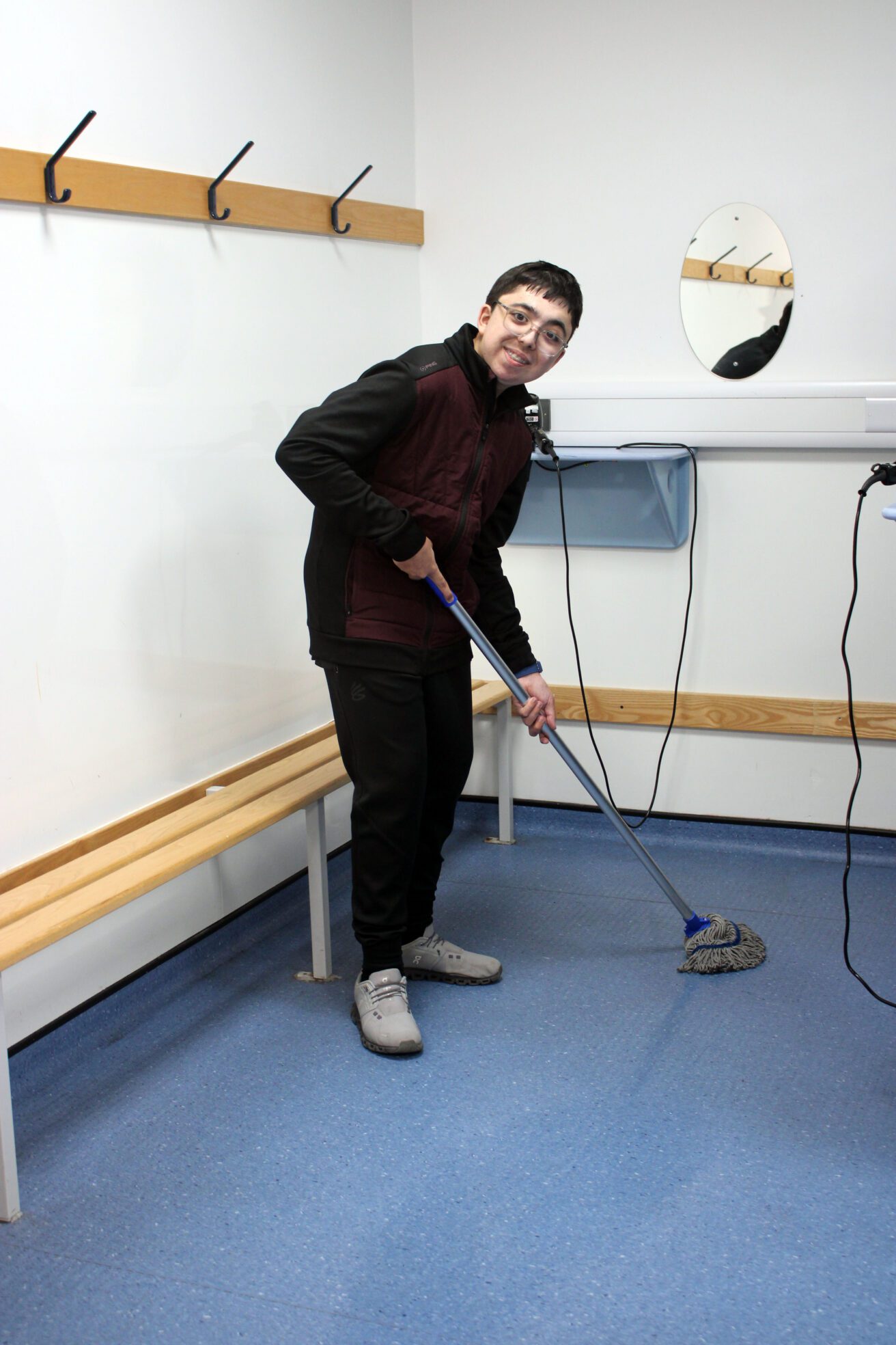Emilio holds the mop the floor with two hands whilst smiling.