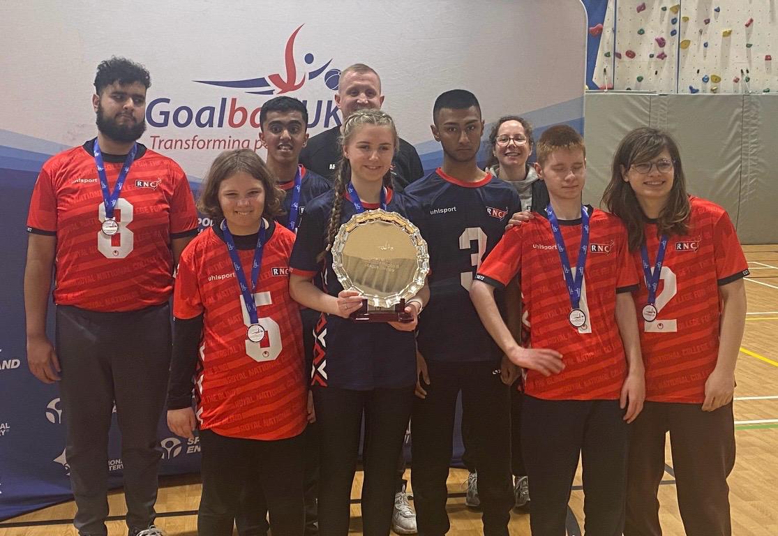 the two teams and their coaches wearing their medals and holding up the silver plate trophy - they are all smiling and standing in front of a Goalball UK banner