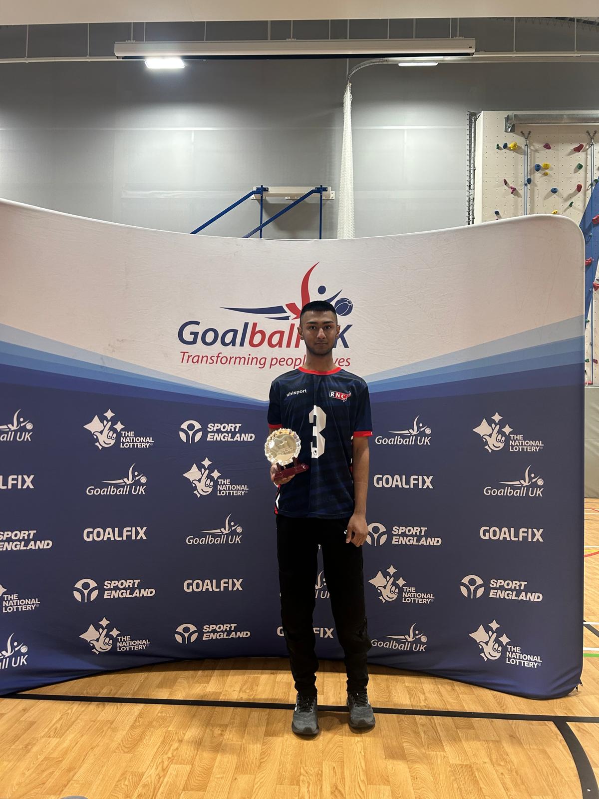 Ishaq in navy blue goalball kit stands in front of a Goalball UK banner holding his award uo