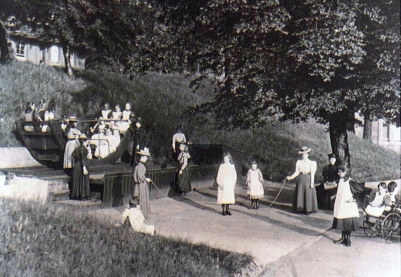 An old photograph showing teachers and students in Victorian outfits. Some are sitting in a seesaw, others are playing skipping rope, and some are sitting down.