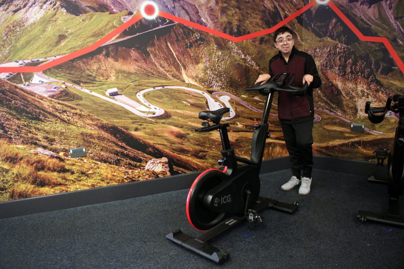 Emilio in the spin studio there's a giant mural behind him of a cycle route going across mountains. Emilio is putting a spin bike.