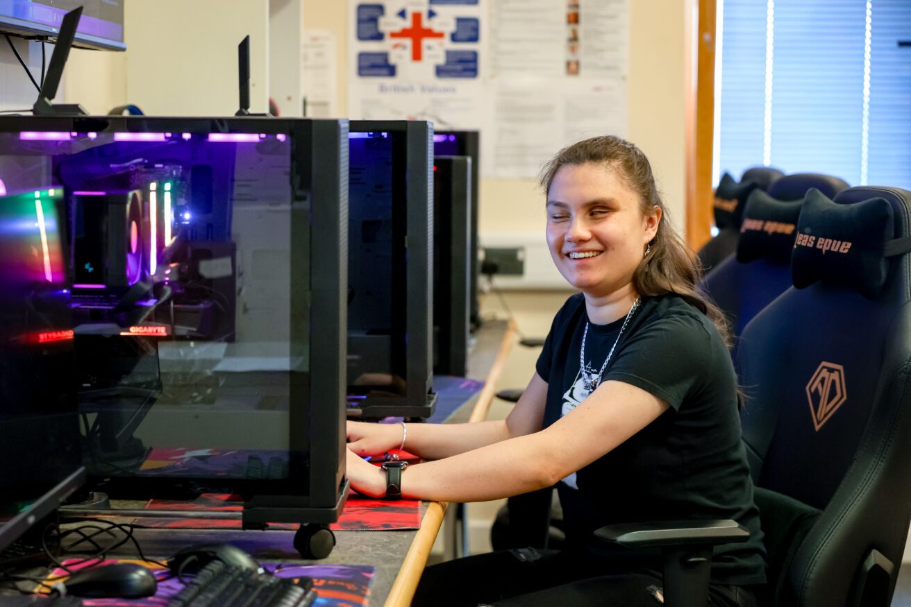 A student sitting at a bank of brightly coloured computers - her hands are on the keyboard and she's looking at the camera smiling