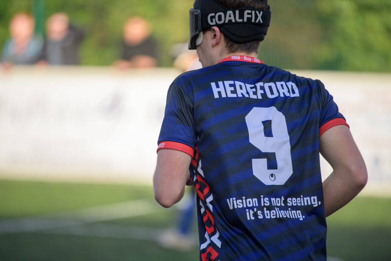 the back of a blind footballer wearing a blue strip and eye shades. He is wearing number 9 and the back of the shirt also reads: 'Hereford Vision is not seeing, it's believing'