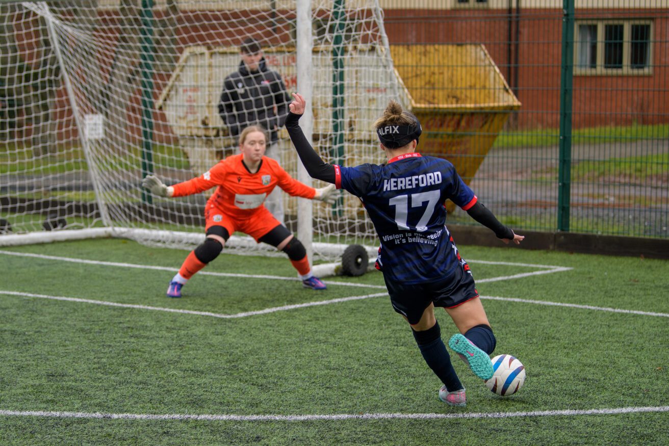 A female football player about to take a shot on goal. The female goalkeeper in orange strip is spreading herself ready for impact