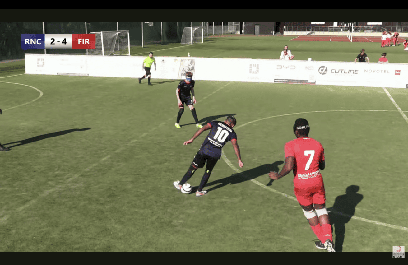 lind football match in progress on an outdoor artificial turf pitch. In the center, a player wearing a dark blue jersey with the number 10 and "HEREFORD" printed on the back controls the ball while wearing eye patches or blindfolds. Nearby, a player in a bright red kit with the number 7 faces toward the play. Another player in dark kit and blindfolds stands in the background alongside a referee in a yellow shirt.