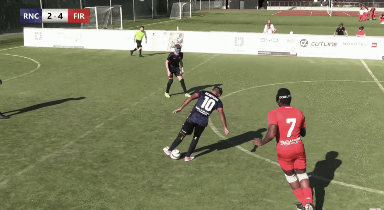 lind football match in progress on an outdoor artificial turf pitch. In the center, a player wearing a dark blue jersey with the number 10 and "HEREFORD" printed on the back controls the ball while wearing eye patches or blindfolds. Nearby, a player in a bright red kit with the number 7 faces toward the play. Another player in dark kit and blindfolds stands in the background alongside a referee in a yellow shirt.