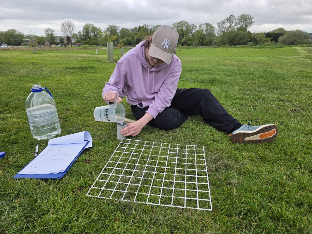 A students sits in a field with a wire grid in front of him - he is pouring water into a beaker and a pad and large bottle of water are to his left