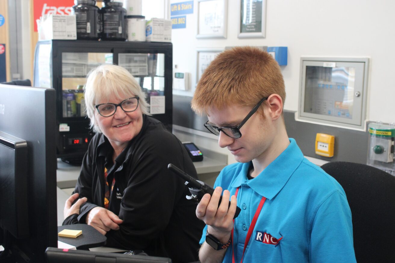 A business student working on thePoint4 reception. He is speaking into a walkie talkie as a receptionist watches on smiling