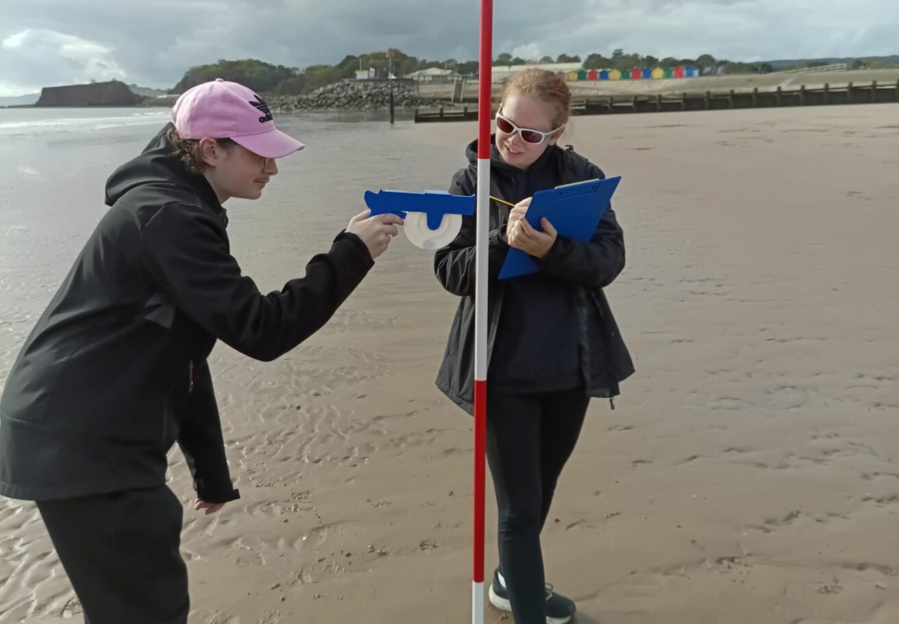 Two students on a beach, one holds a clipboard and the other is using a measuring tool against a pole
