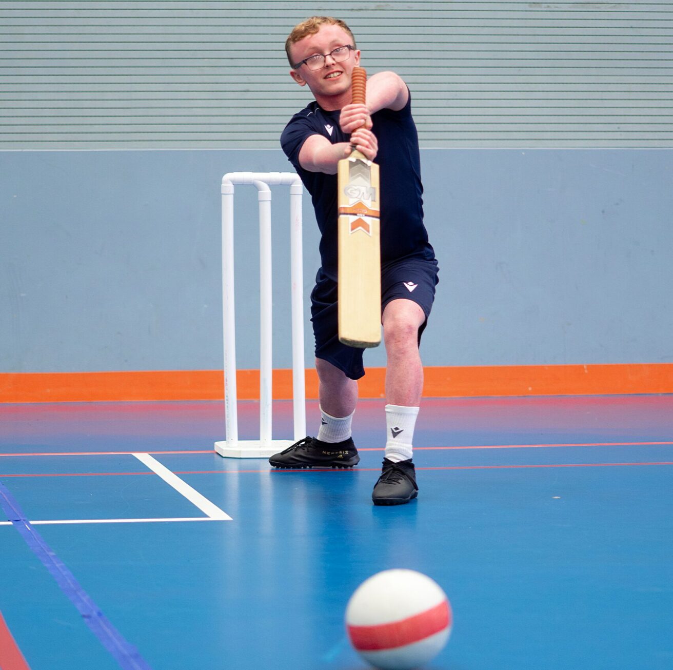 A player in the sports hall stands in front of the stumps about to hit the ball with a cricket bat