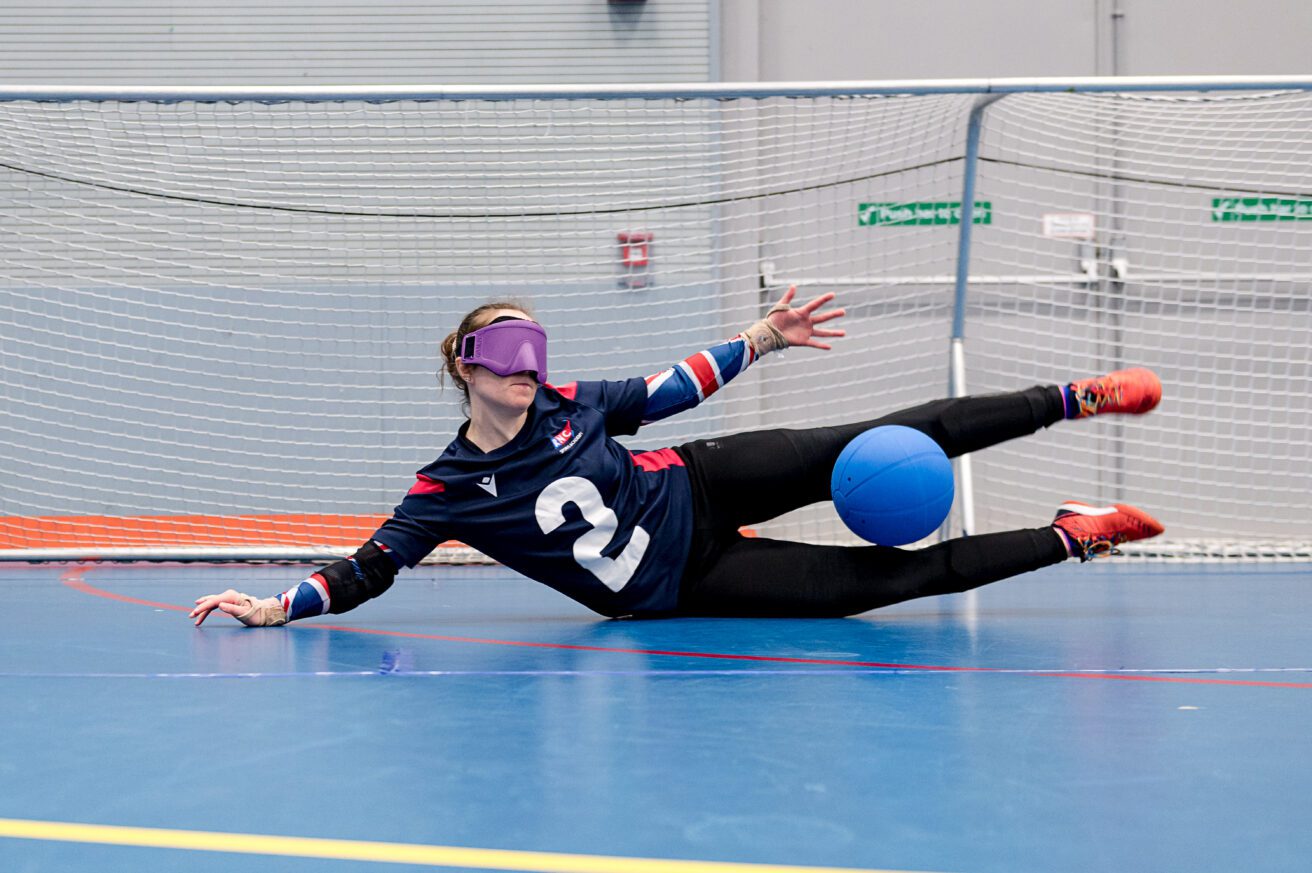 A goalball player in navy kit and eyeshades defending goal she has thrown her body across goal to trap the ball with her legs