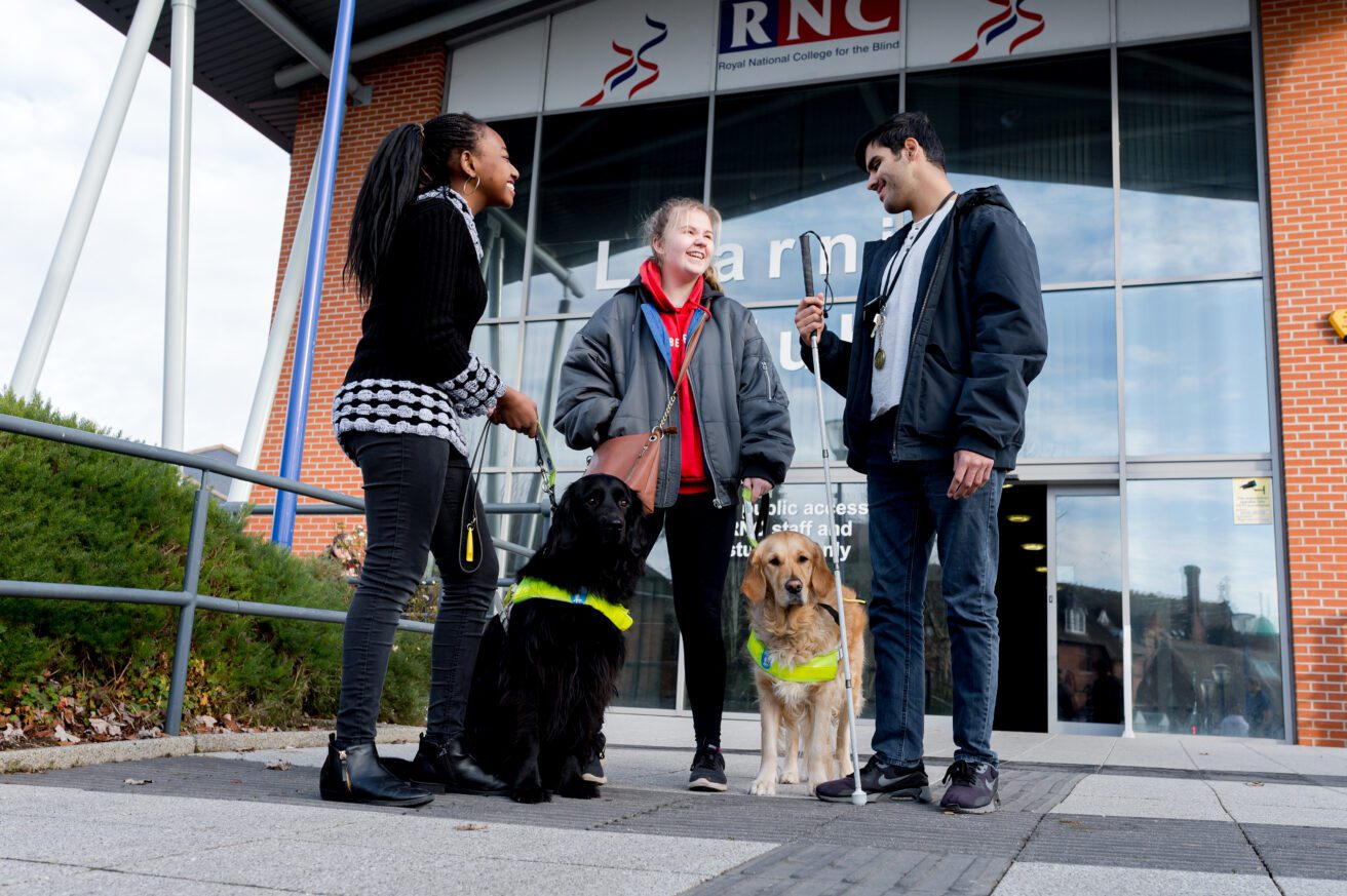 Three students and two guide dogs standing in front of the entrance to the College's learning hub