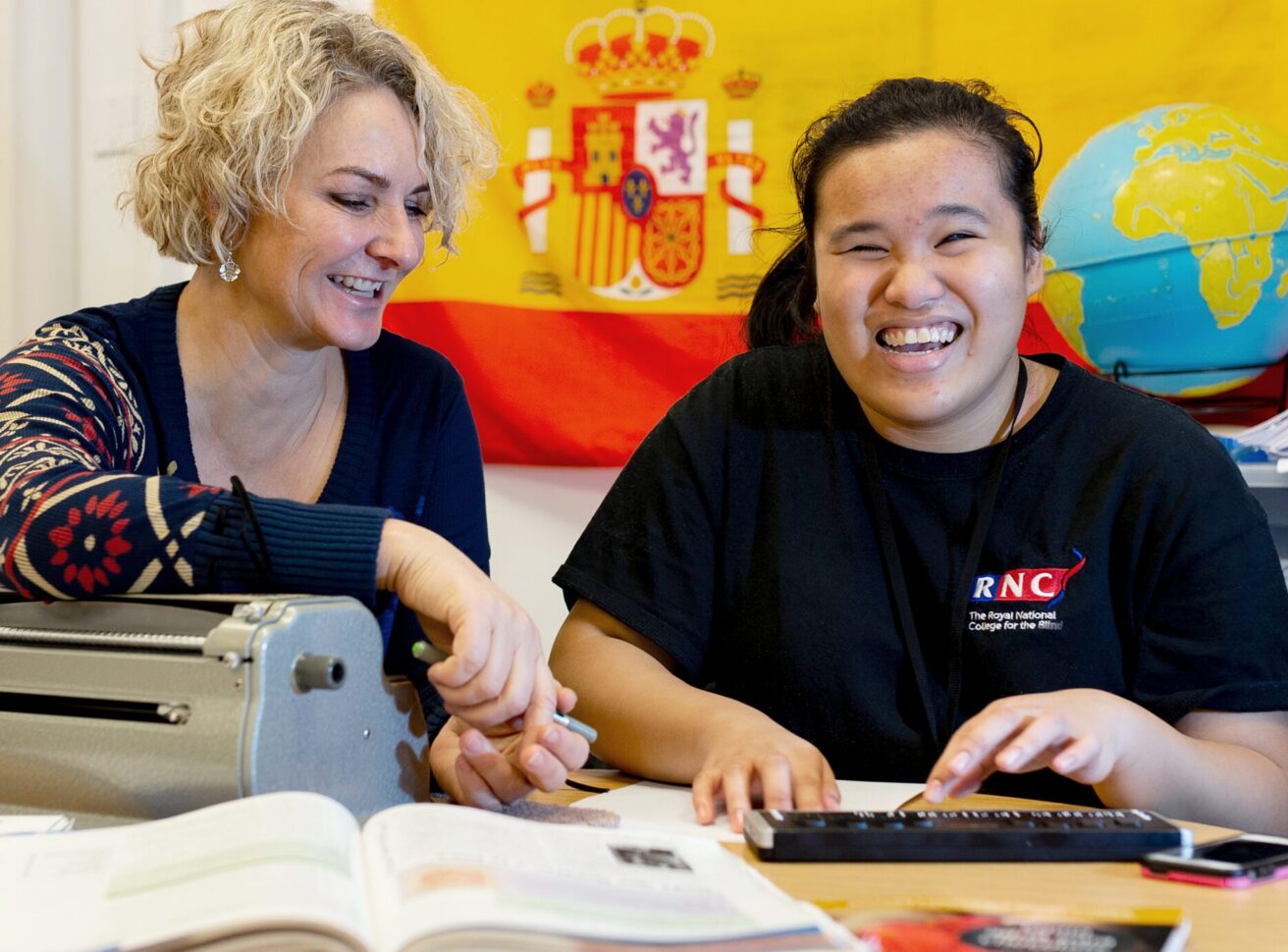 A student using a braille note in a Spanish class. A teacger sits next to her leaning on Perkins brailler machine. There is a large Spanish flag displayed on the wall behind them and a globe sits on a shelf behind the student who is smiling