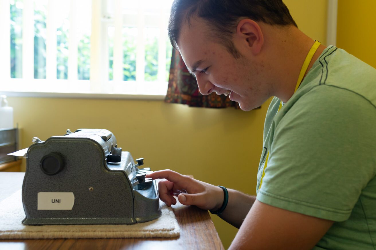 A side view of a smiling student sitting at a table in front of a Perkins Braille machine. He is pressing down on a button with a finger on his right hand