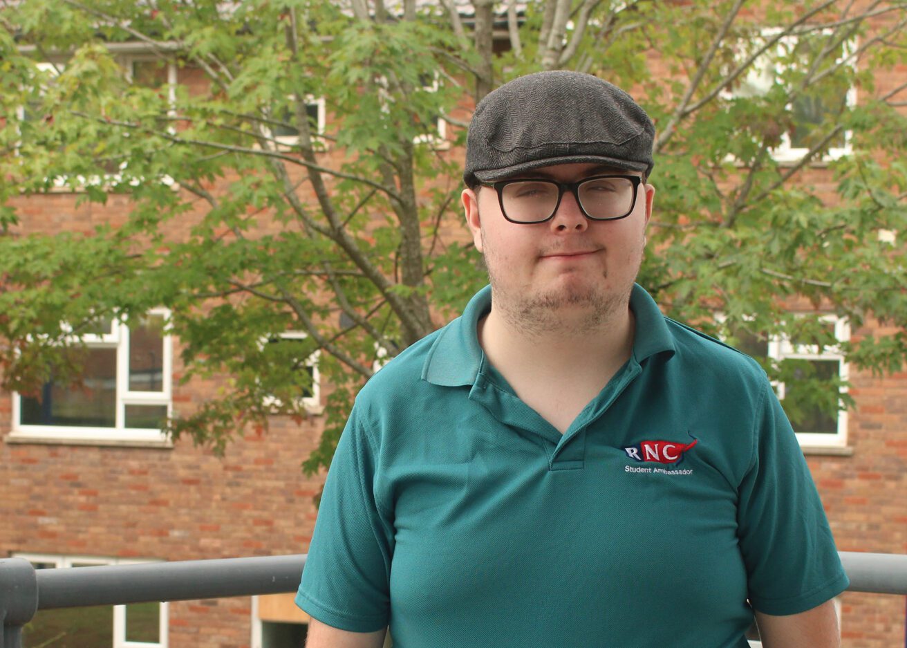 Head and shoulders portrait of Freddie wearing his dark green student ambassador polo shirt and a flat cap