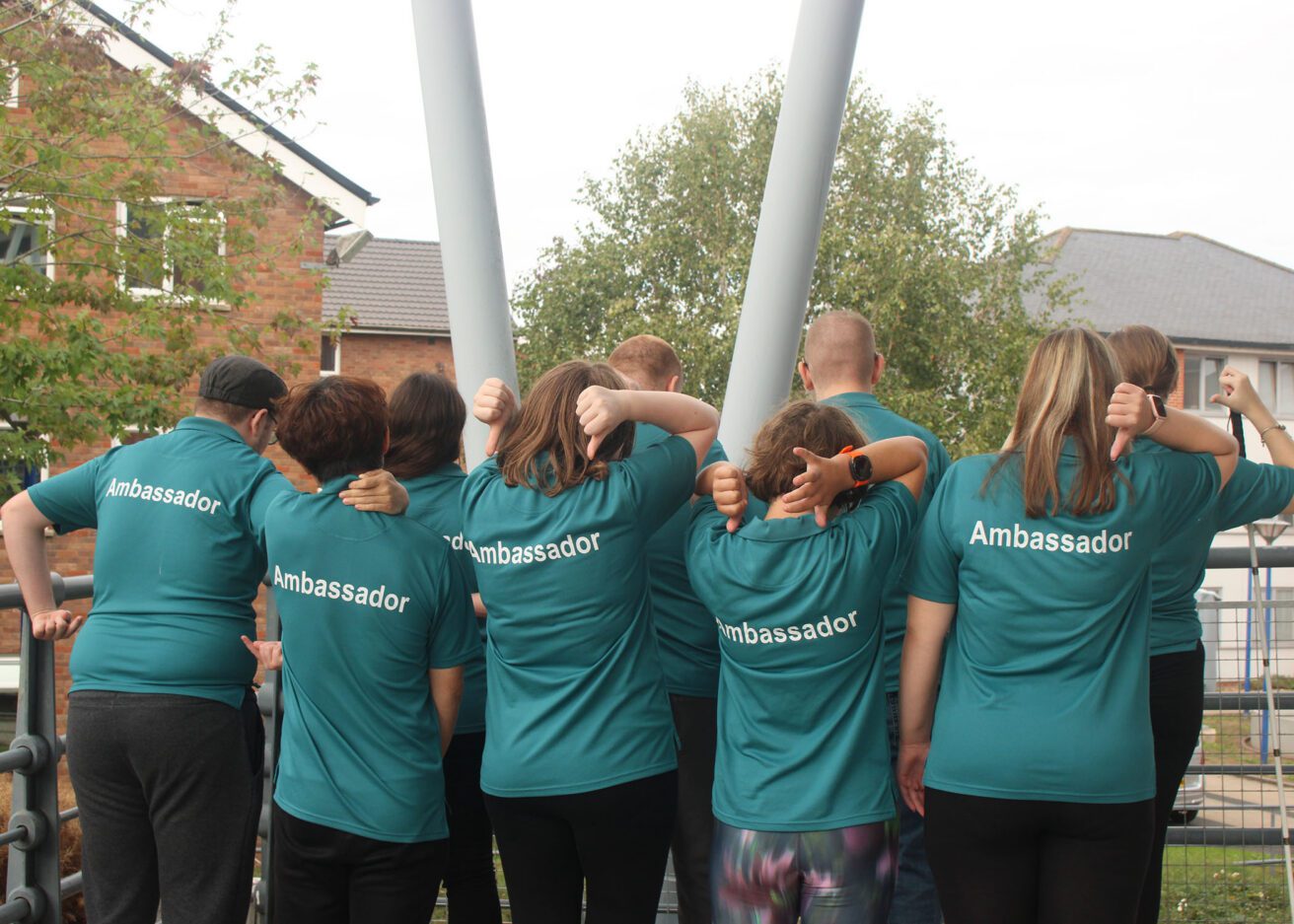 The group of 9 student ambassadors wearing their dark green polo shirts with their backs to camera pointing at the giant writing of ambassador on the backs of their t-shirts