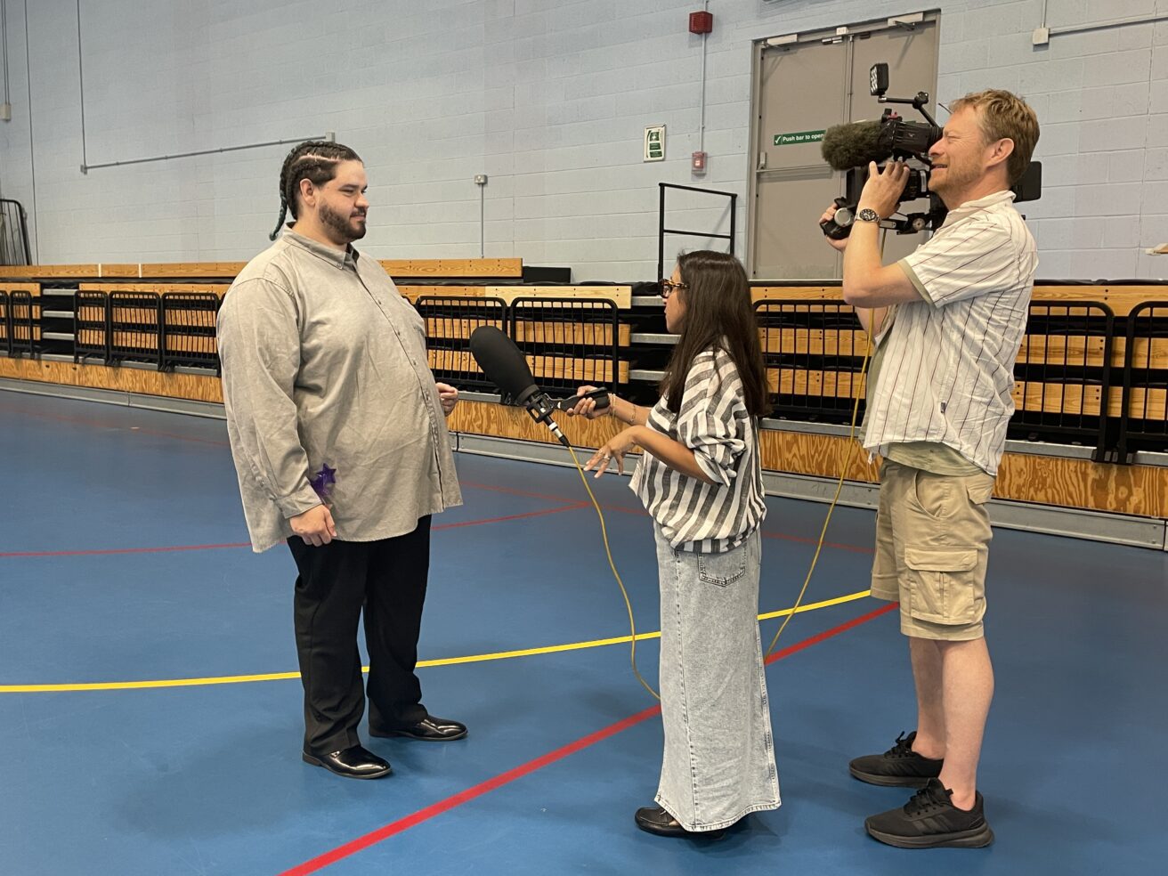 A student talking to a presenter who is holding a microphone towards him. A cameraman is filming their discussion