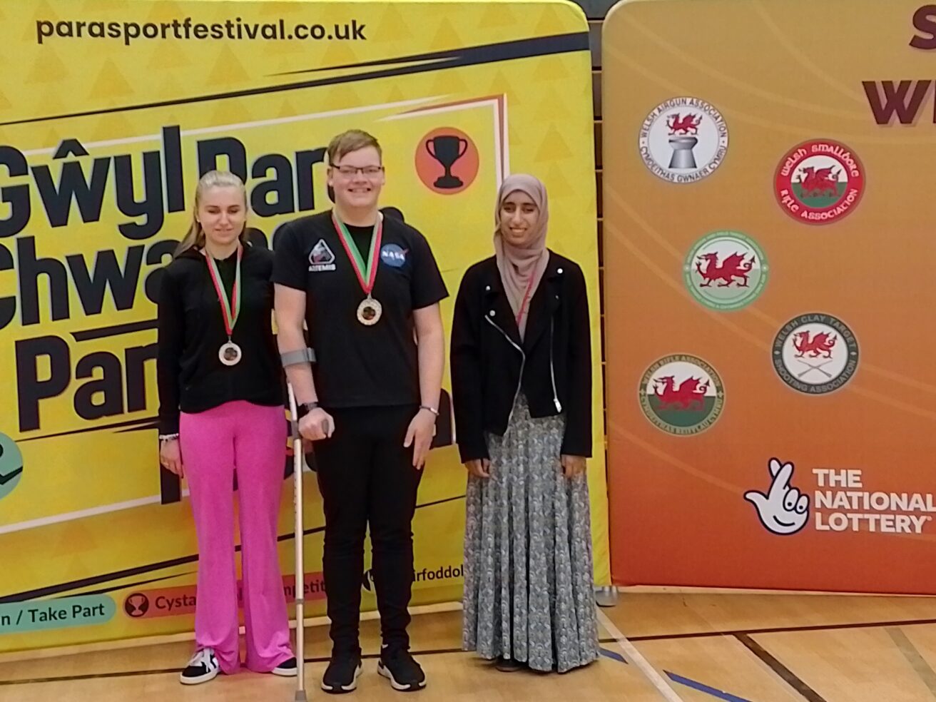 Tegan, Harvey and Humna standing in front of brightly coloured competition promotion boards wearing their medals