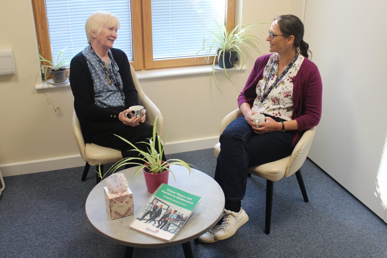 Two SEMH coaches sitting opposite each other chatting and both holding coffee cups. There is a plant a book and a box of tissues in front of them on the small round coffee table