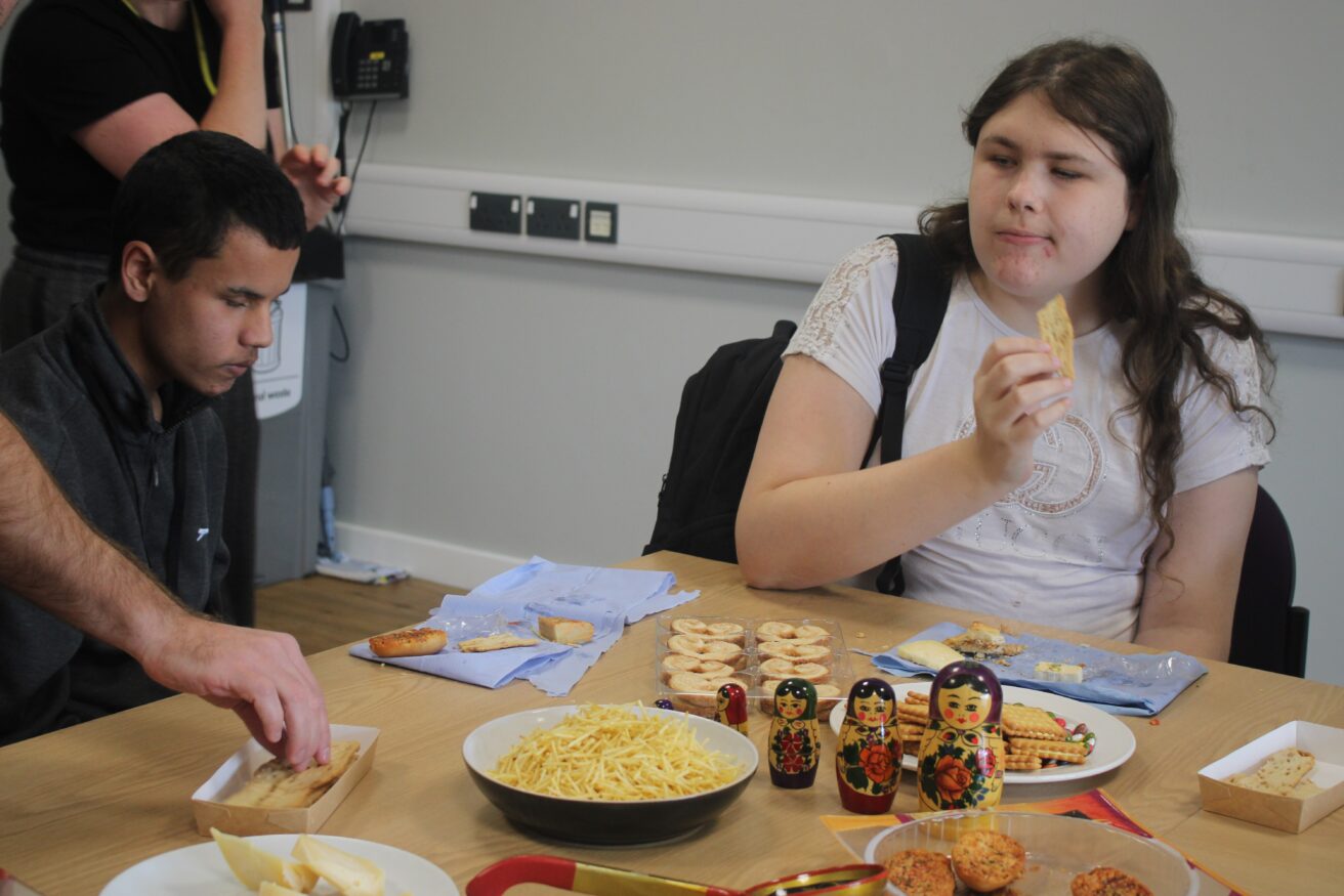 Students seated at a table sampling foods during the celebration. Traditional Russian Matryoshka nesting dolls, crackers, and bowls of snacks are arranged in front of them.