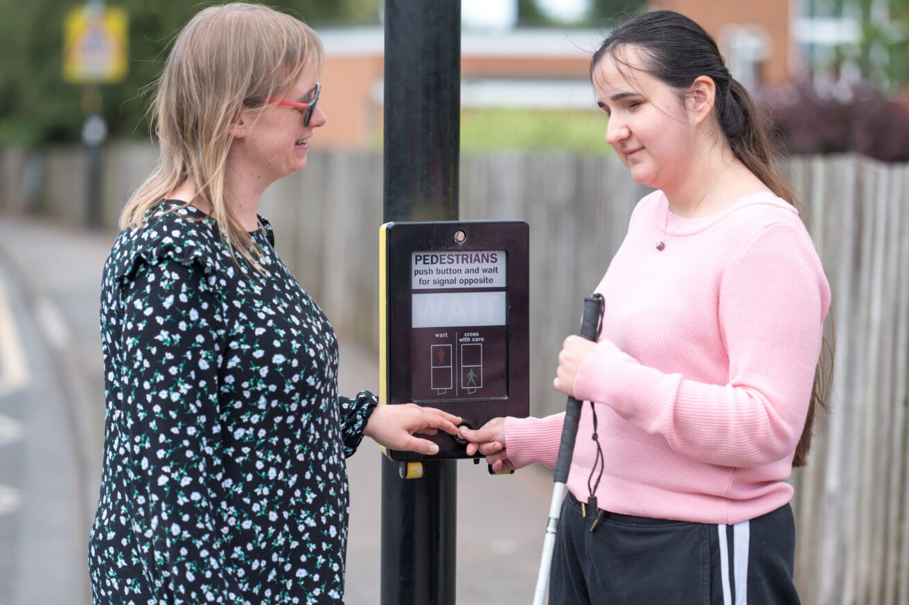 a female teacher and a female student standing next to a road crossing in a mobility lesson - the student is about to press the button