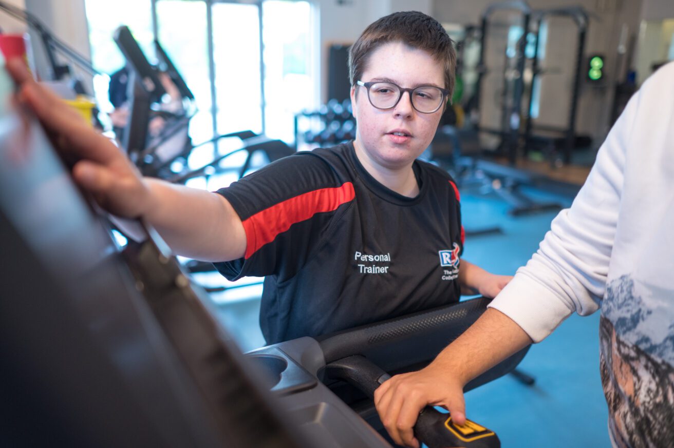 A trainee personal trainer has her hands on the controls of a treadmill showing a client out of shot how to use the apparatus