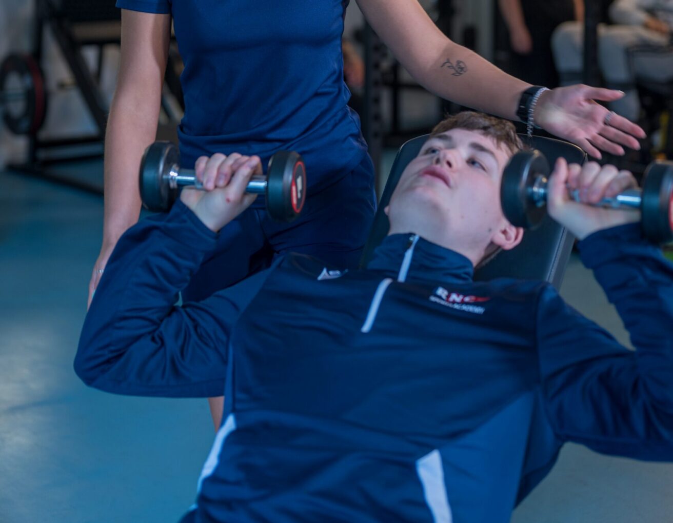 A student lying on a bench with a dumbbell in each hand about to raise them above is head. The arms of a member of staff out of shot are ensuring the activity is being undertaken safely