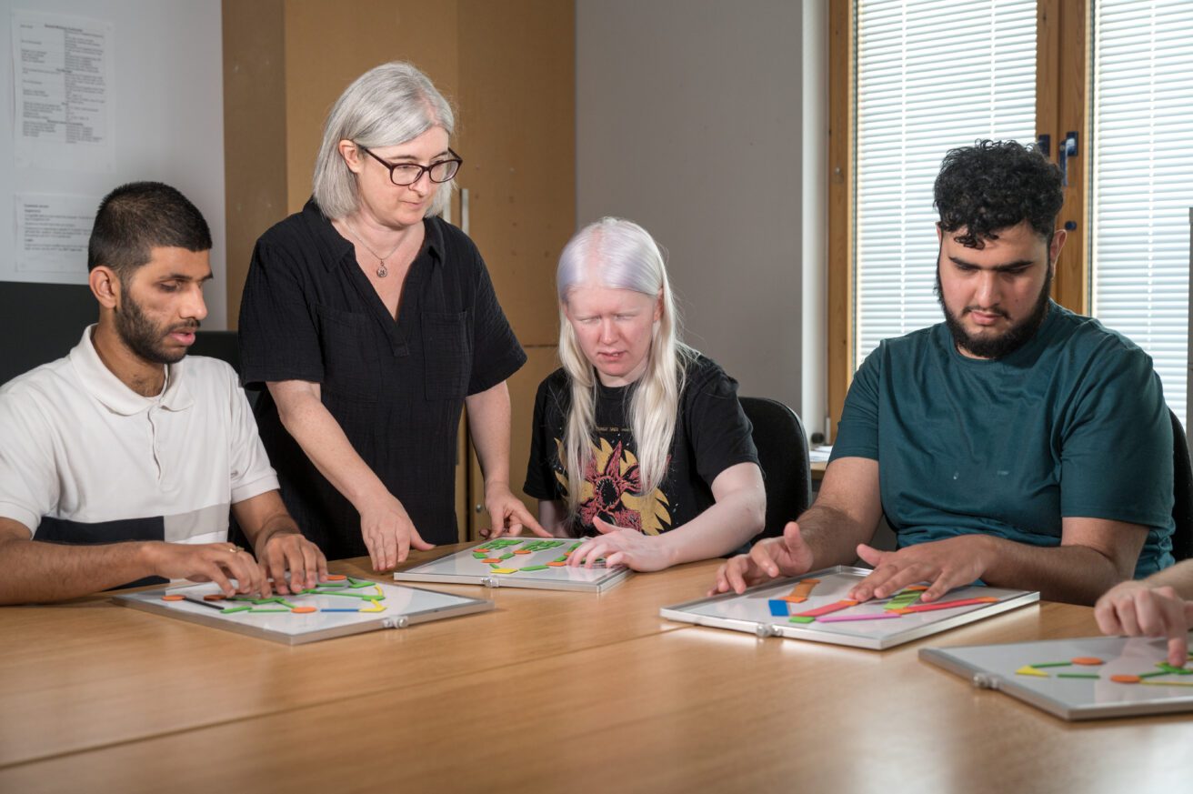 3 students sitting at a table in a classroom using some tactile objects to understand a company structure chart. The teacher stands inbetween them explaining