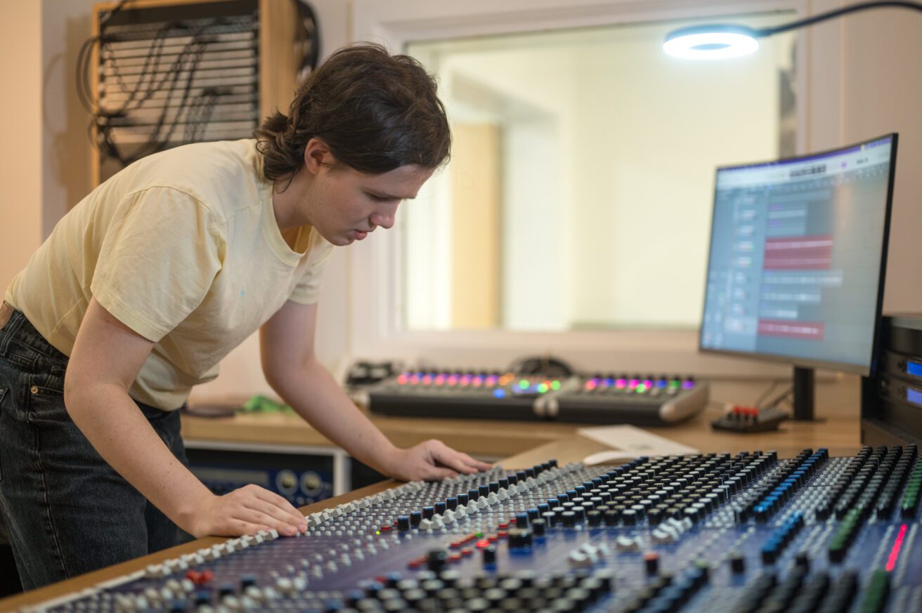 A student working on the mixing desk in the audio media suite. She is looking down at the desk with her hands placed on the edge of the desk