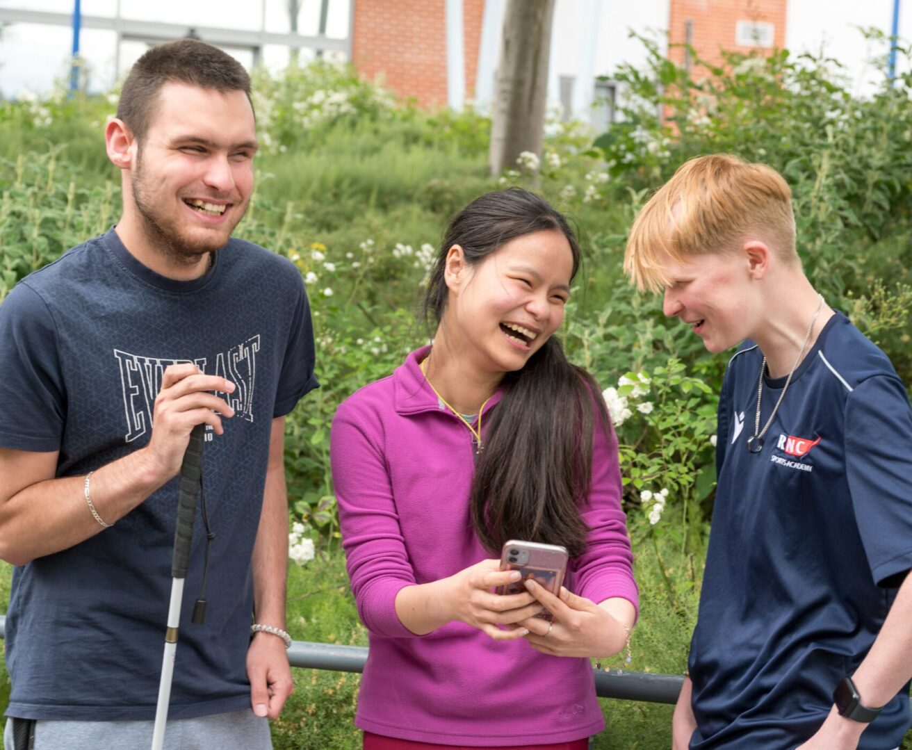 3 students standing outside socialising - the student in the centre is holding a mobile phone and they are all laughing