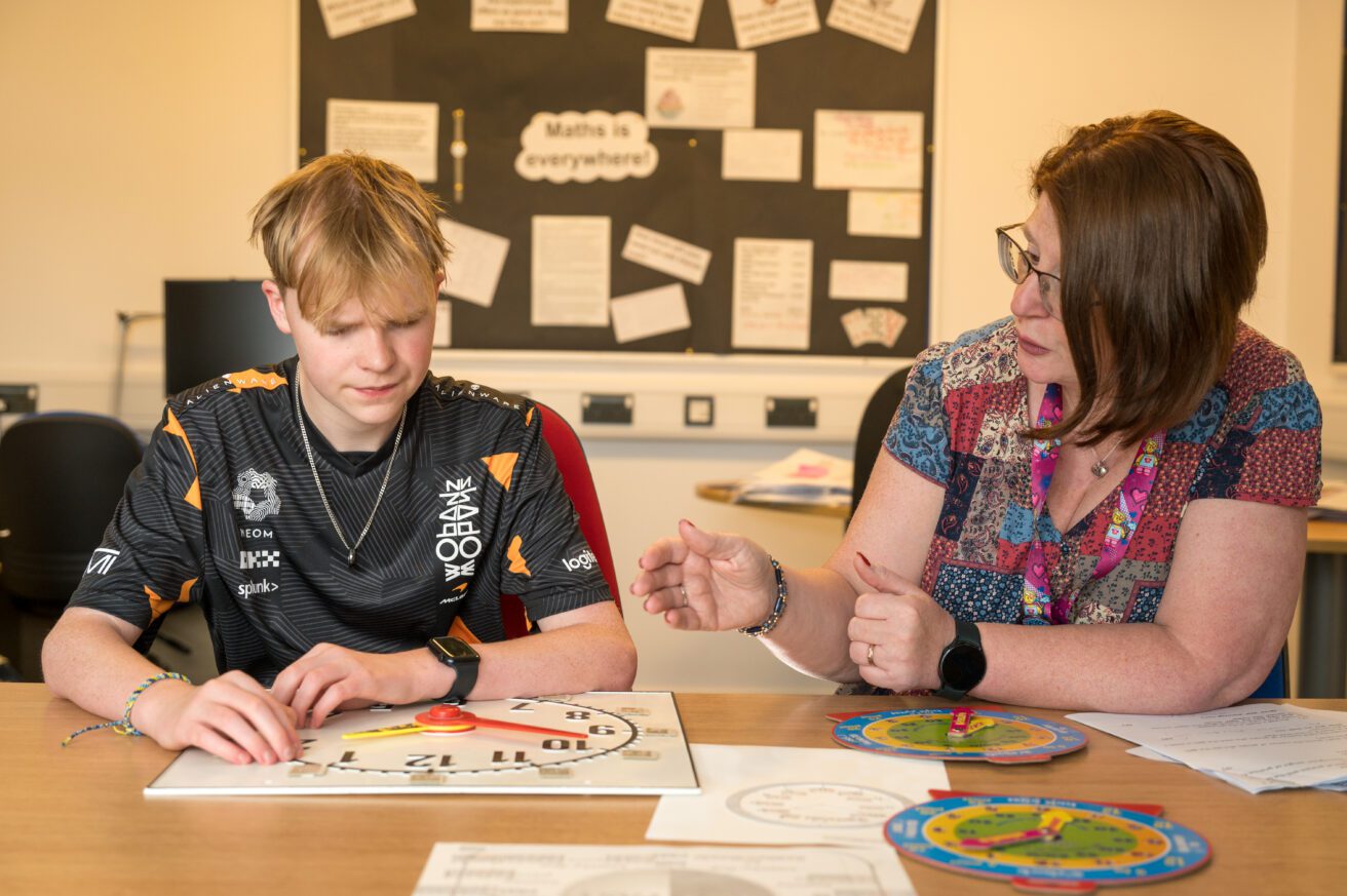 A student sits next to a reacher and they are working on a clock face lying on the desk in front of them