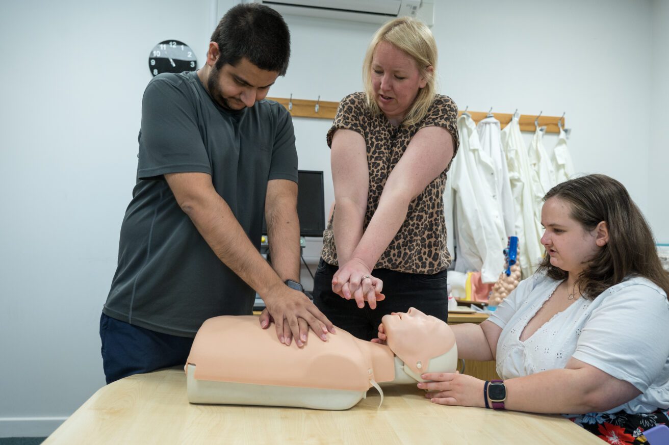 A teacher demonstrates how to position hands for CPR. A Resusci Anne manekin is on the desk