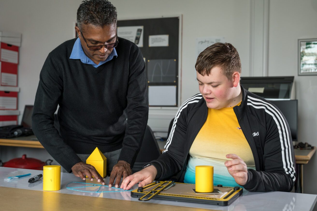 A Maths class - a student sits at a desk with various 3D shapes and a tactipad in front of him. The teacher stands to his right and is guiding him through a tactile shape on the desk