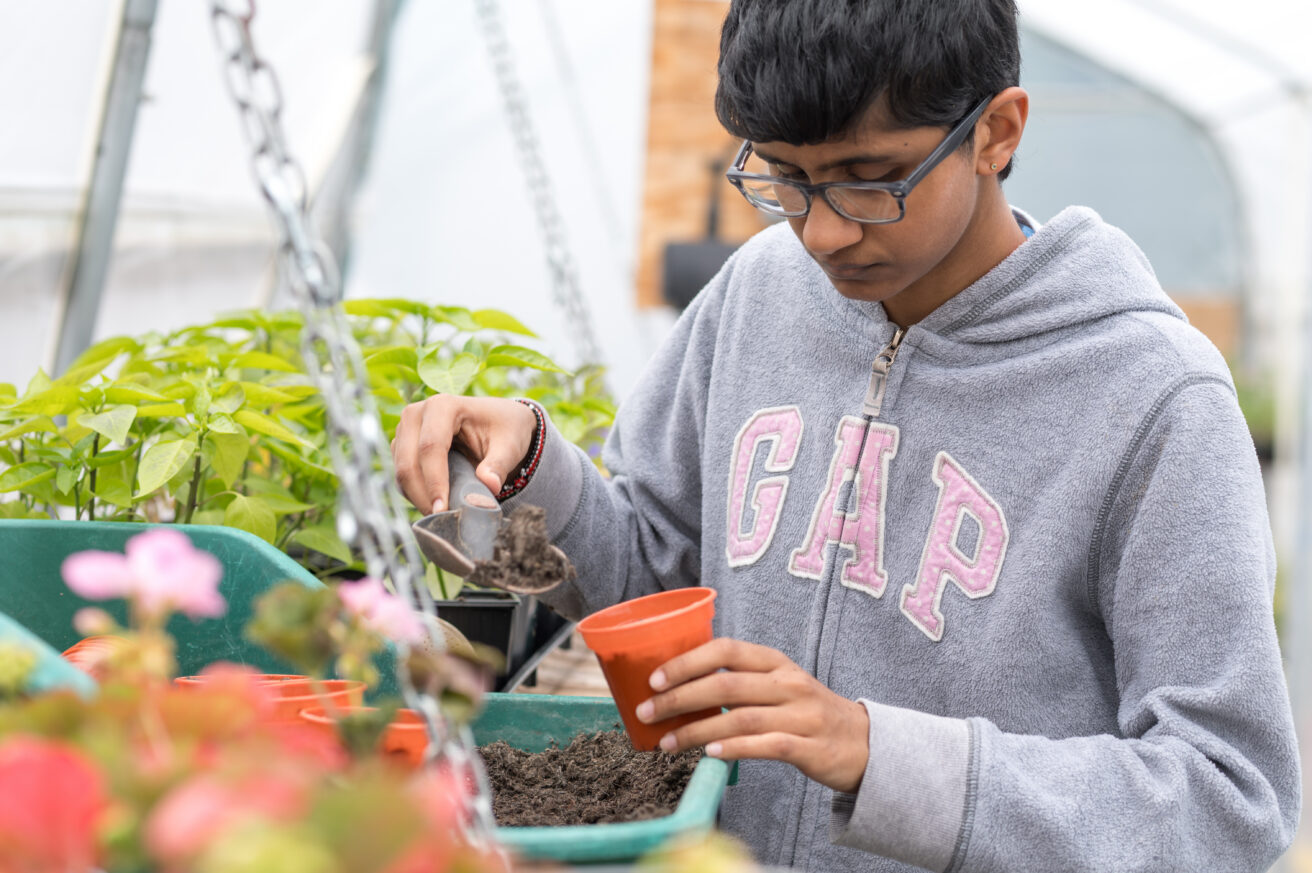 a student standing in a polytunnel using a trowel to fill a flower pot with compost